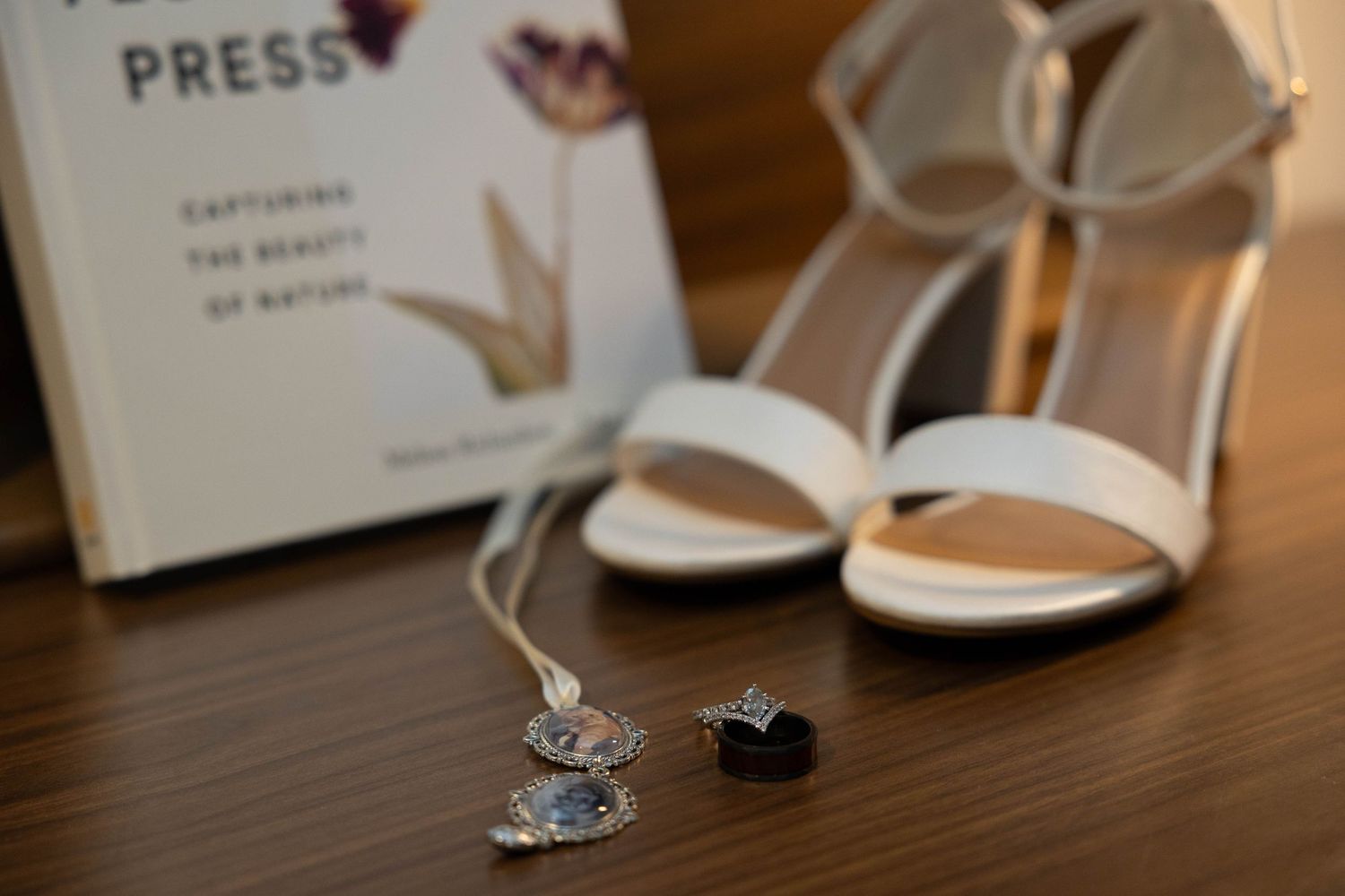 Wedding details flat lay with white heels, rings, and necklace on wood surface.