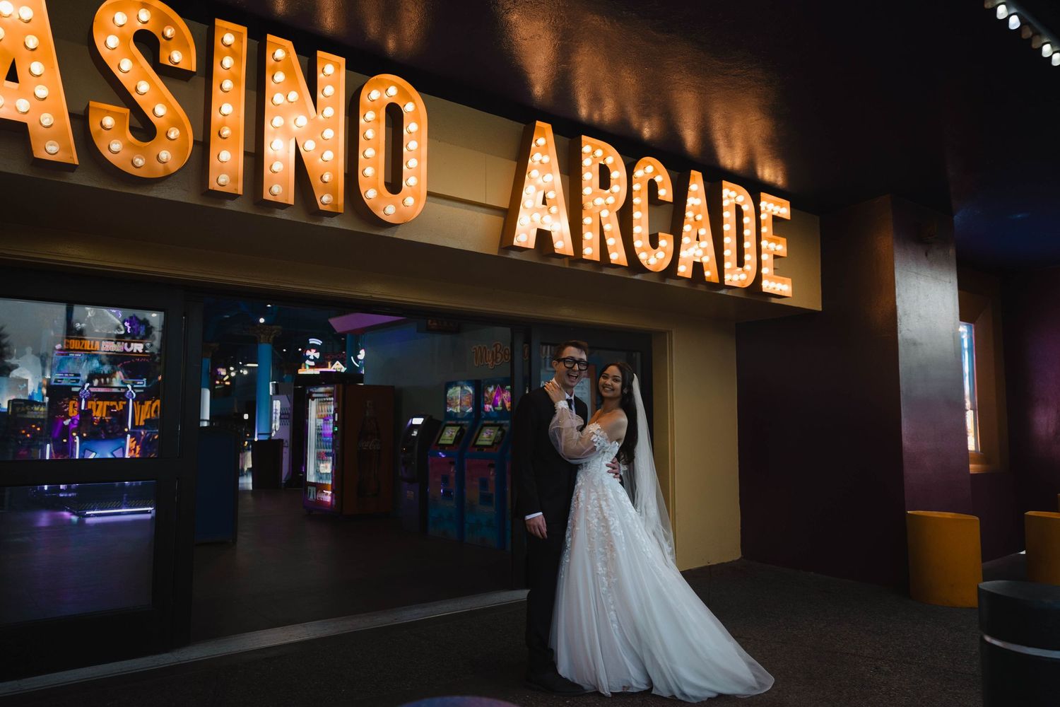 Newlywed couple embracing in wedding attire outside a Casino Arcade entrance with marquee lights.