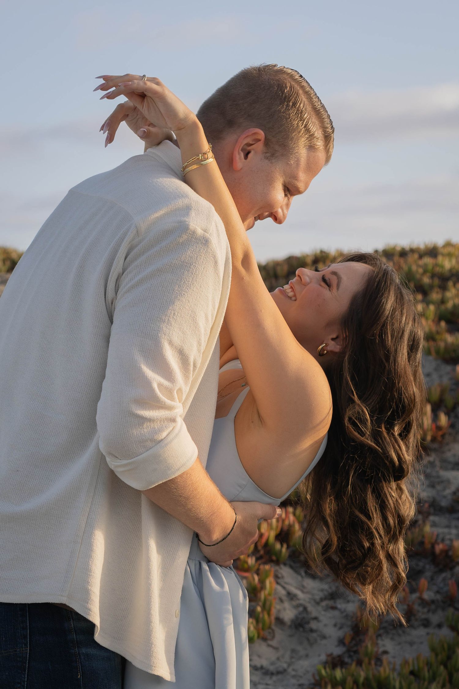 A couple shares a romantic moment outdoors, the woman laughing as the man dips her near sandy dunes at sunset. Coronado beach couples photos