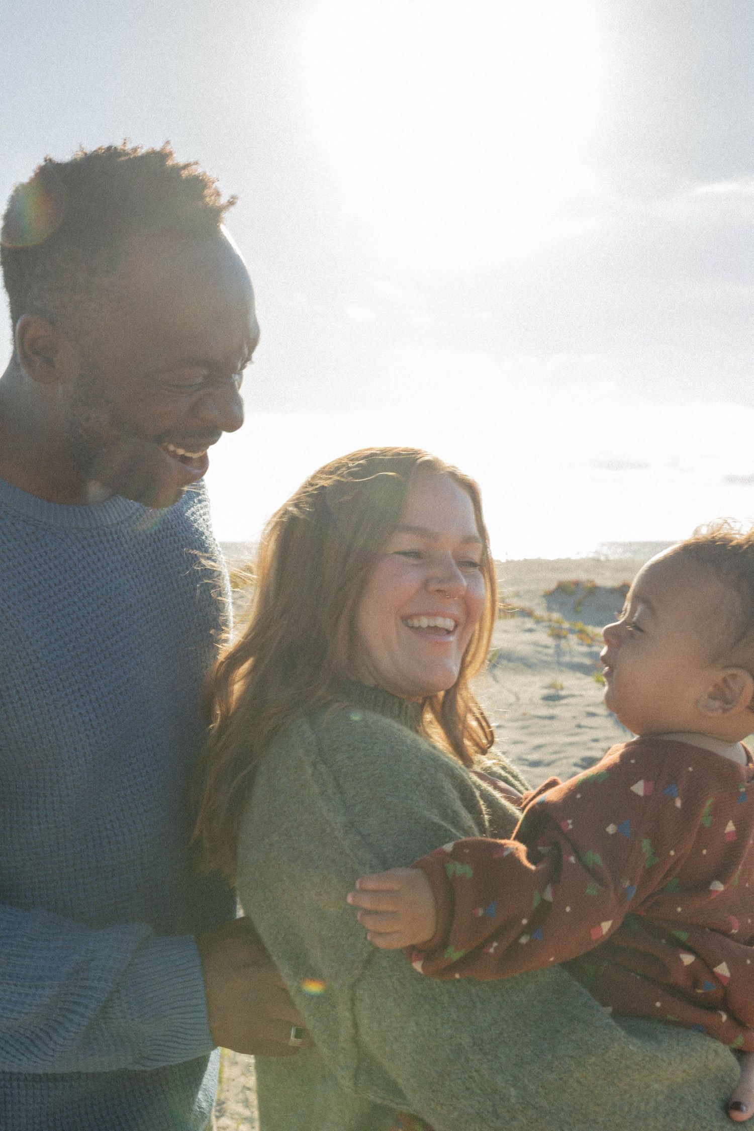 Family laughing together outdoors on a sunny beach, parents holding their baby child. Coronado beach family photographer.