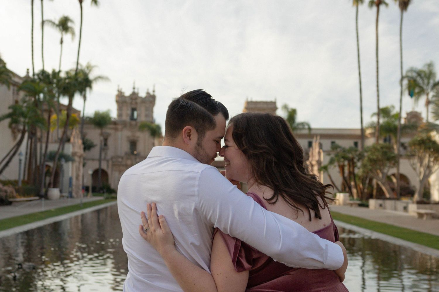 Couple embracing romantically near a reflecting pool with palm trees and historic architecture at Balboa Park, San Diego. Couples photograph