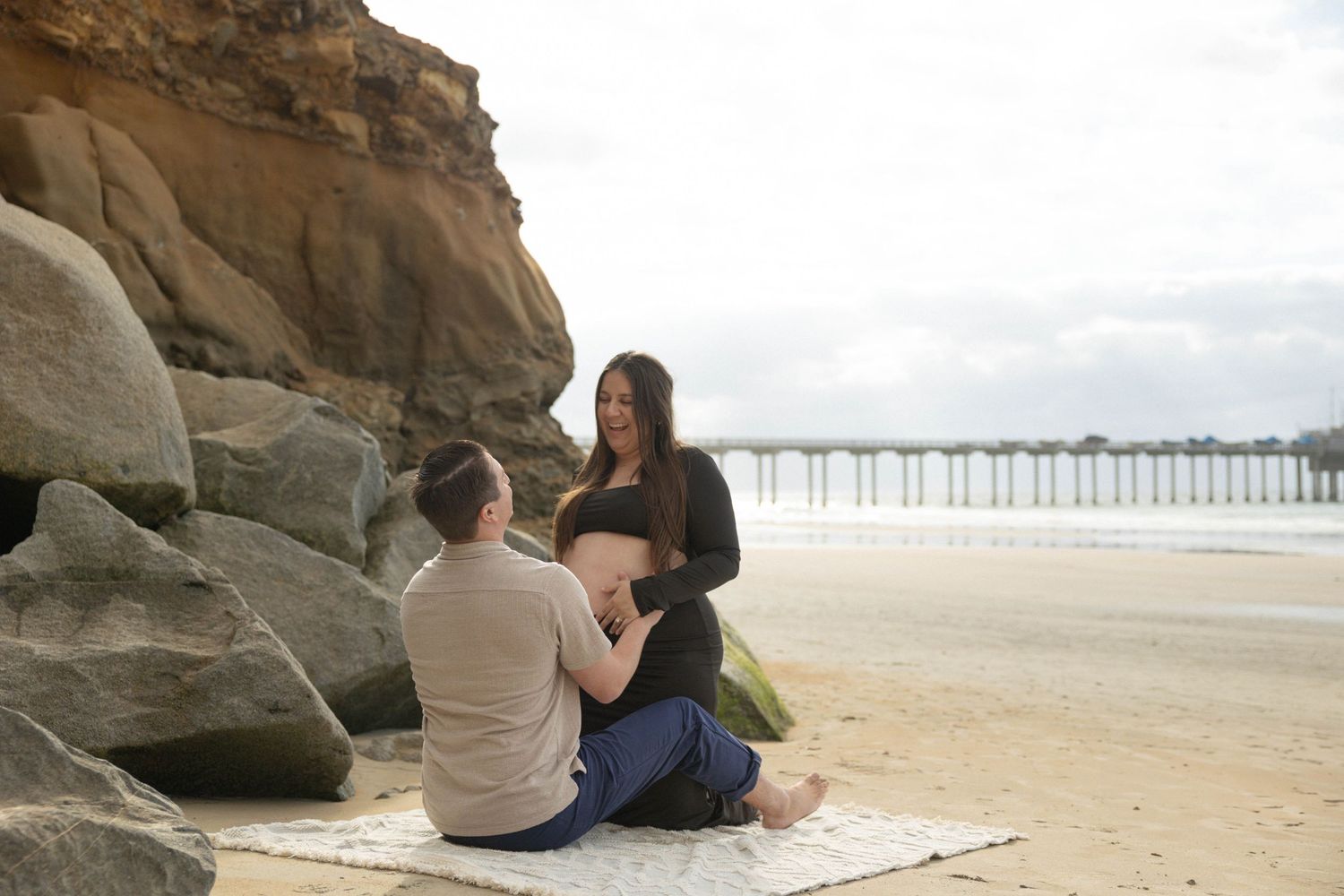 Couple sitting on beach blanket near rocky cliffs with pier in background during maternity photo session. Maternity Photos La Jolla SD.