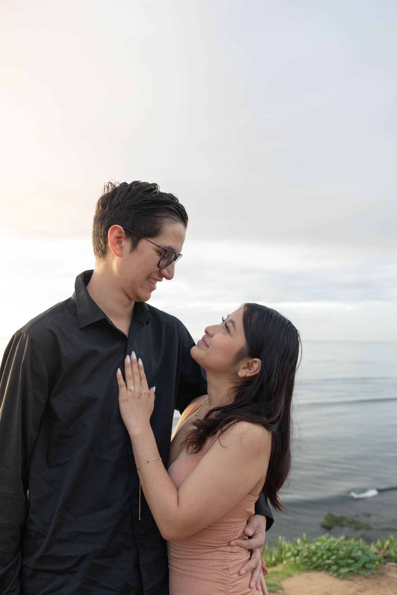 Couple sharing a romantic moment by the ocean, woman in pink dress with engagement ring, man in black shirt. San Diego Engagement Photograph