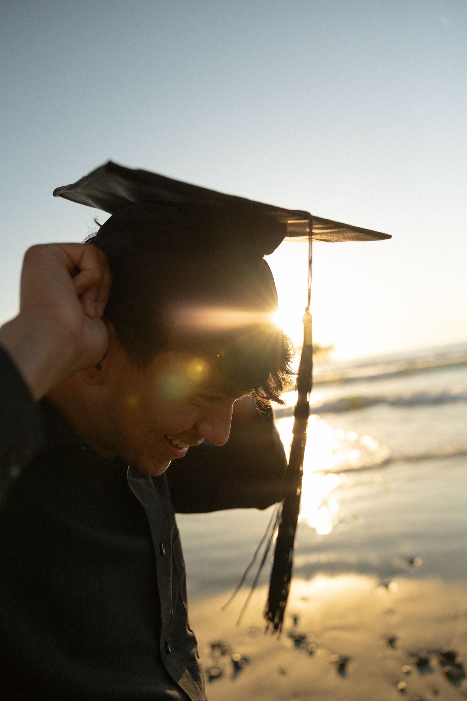Graduate wearing mortarboard cap at sunset beach with golden lens flare reflecting on ocean water.