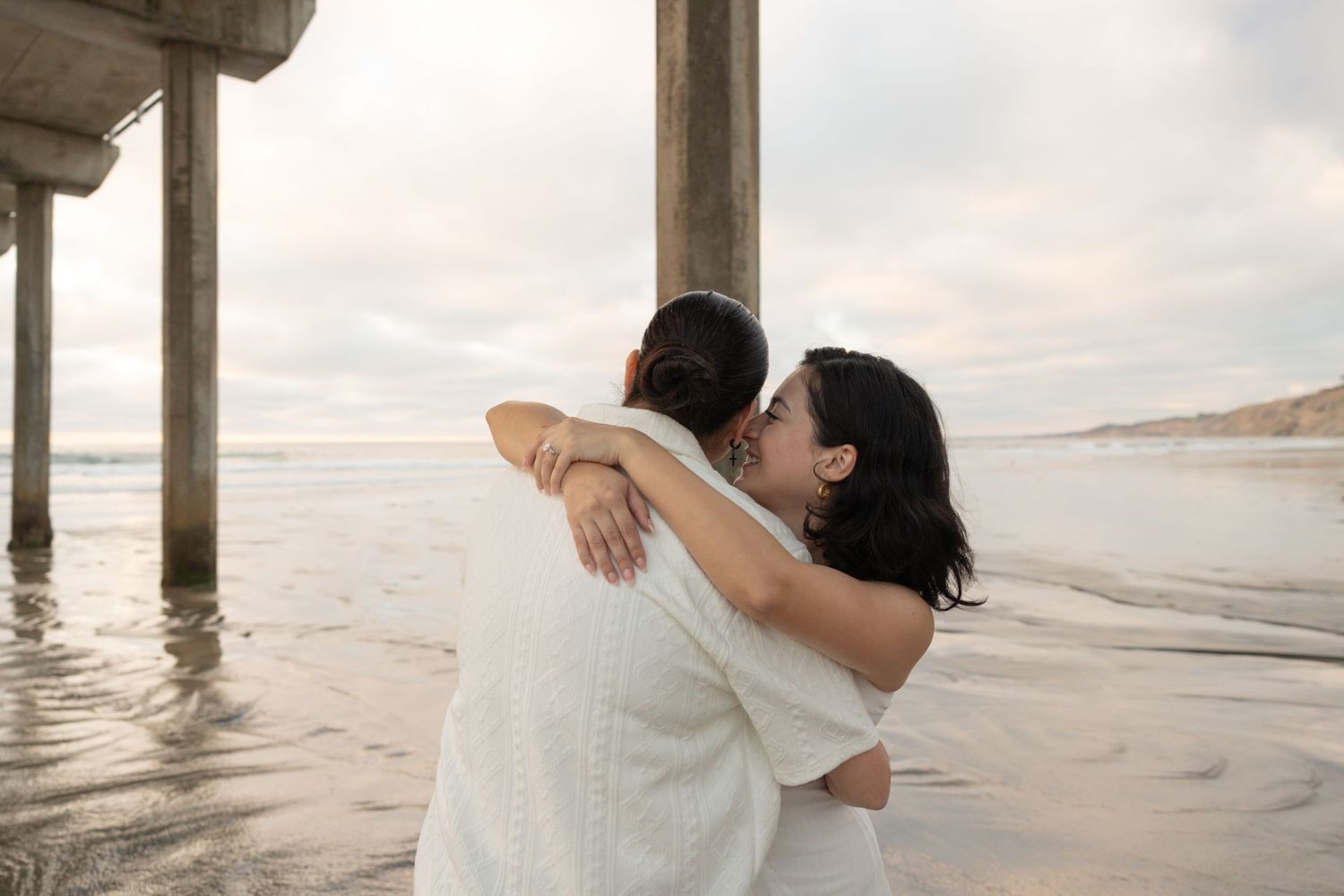 Two women share a tender embrace on a beach near a pier at sunset.