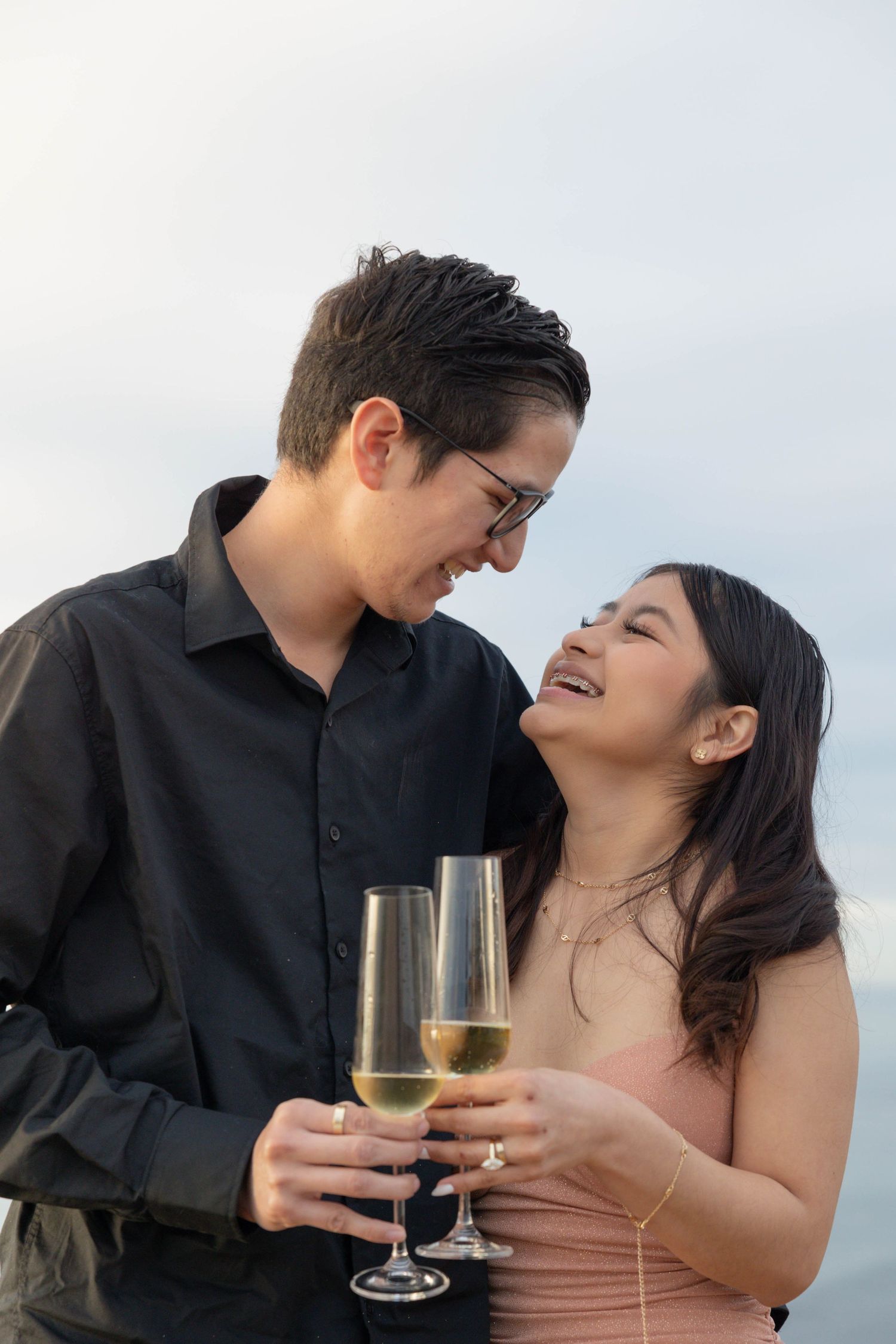 Happy couple toasting with champagne glasses outdoors, smiling at each other in a romantic celebration moment. Engagement Photographer.