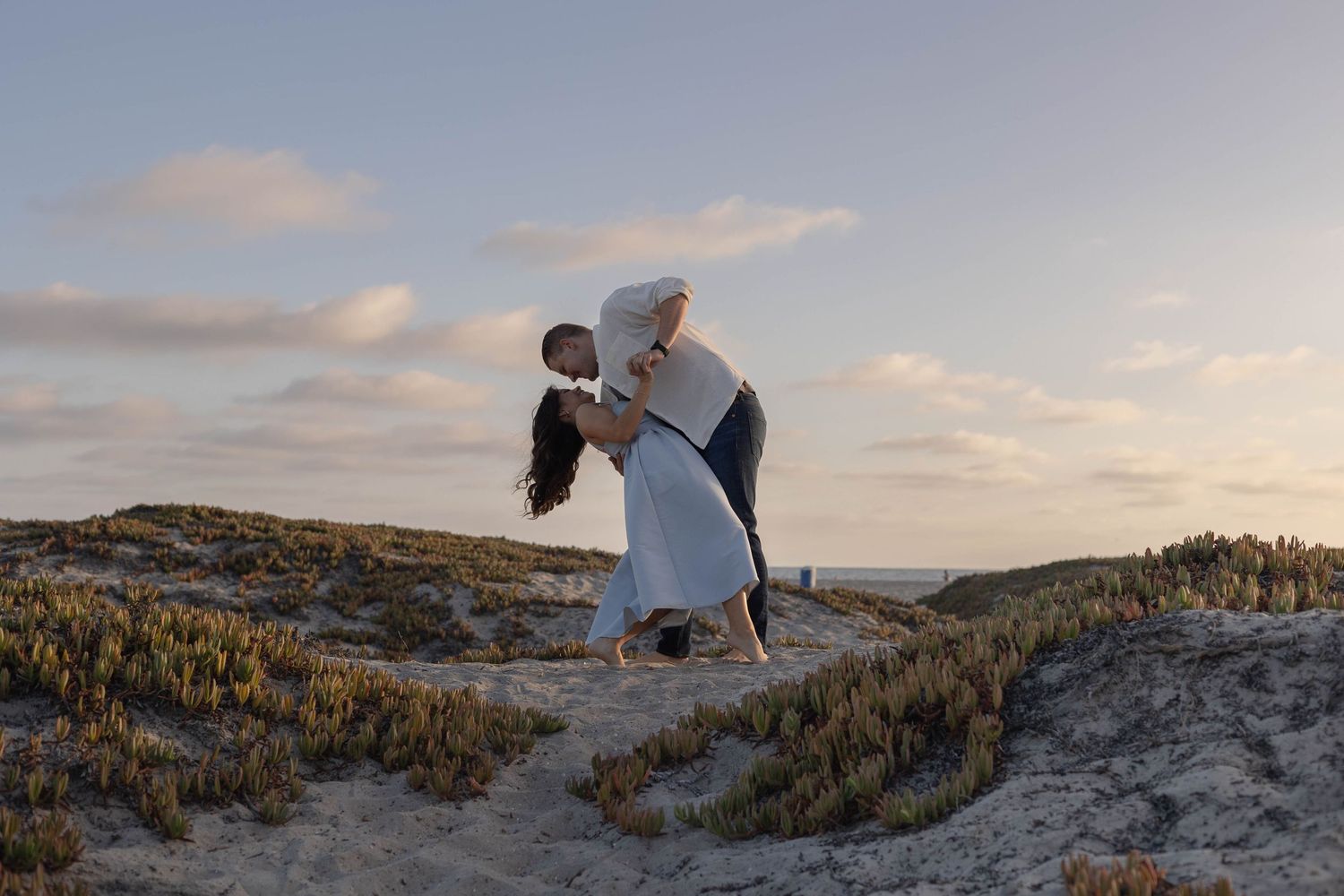 Couple sharing a romantic dip kiss on sandy beach dunes at sunset with soft cloudy sky backdrop.