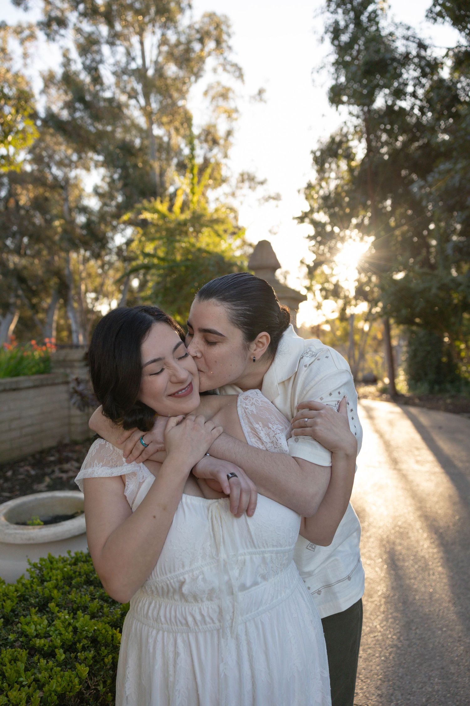 Two women in white dresses share a tender embrace and kiss outdoors, surrounded by lush trees in golden sunlight. Couples Portraits SD
