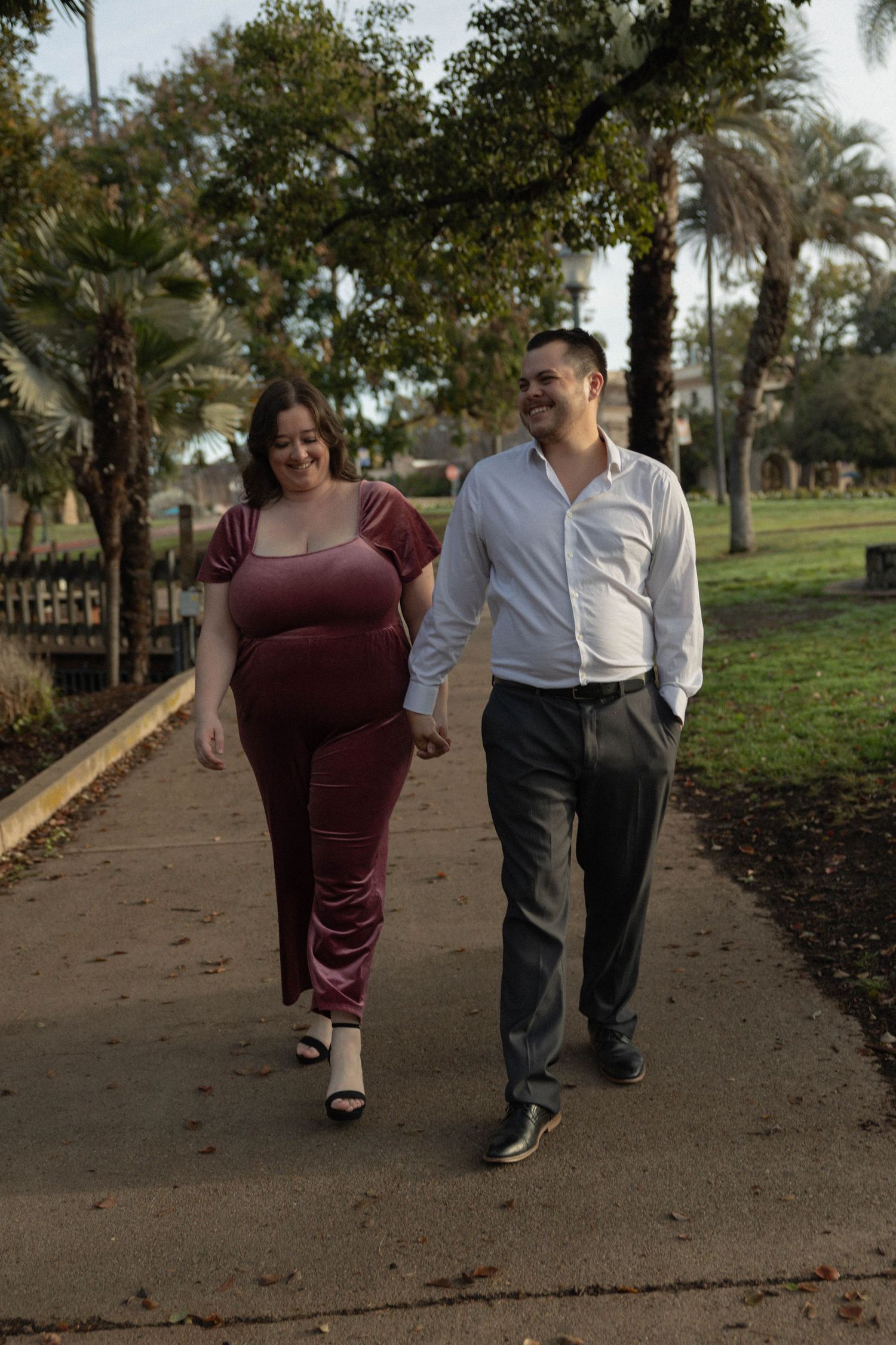 Couple holding hands walking on a park path, woman in burgundy velvet dress and man in white shirt and gray trousers. Couples Portraits SD
