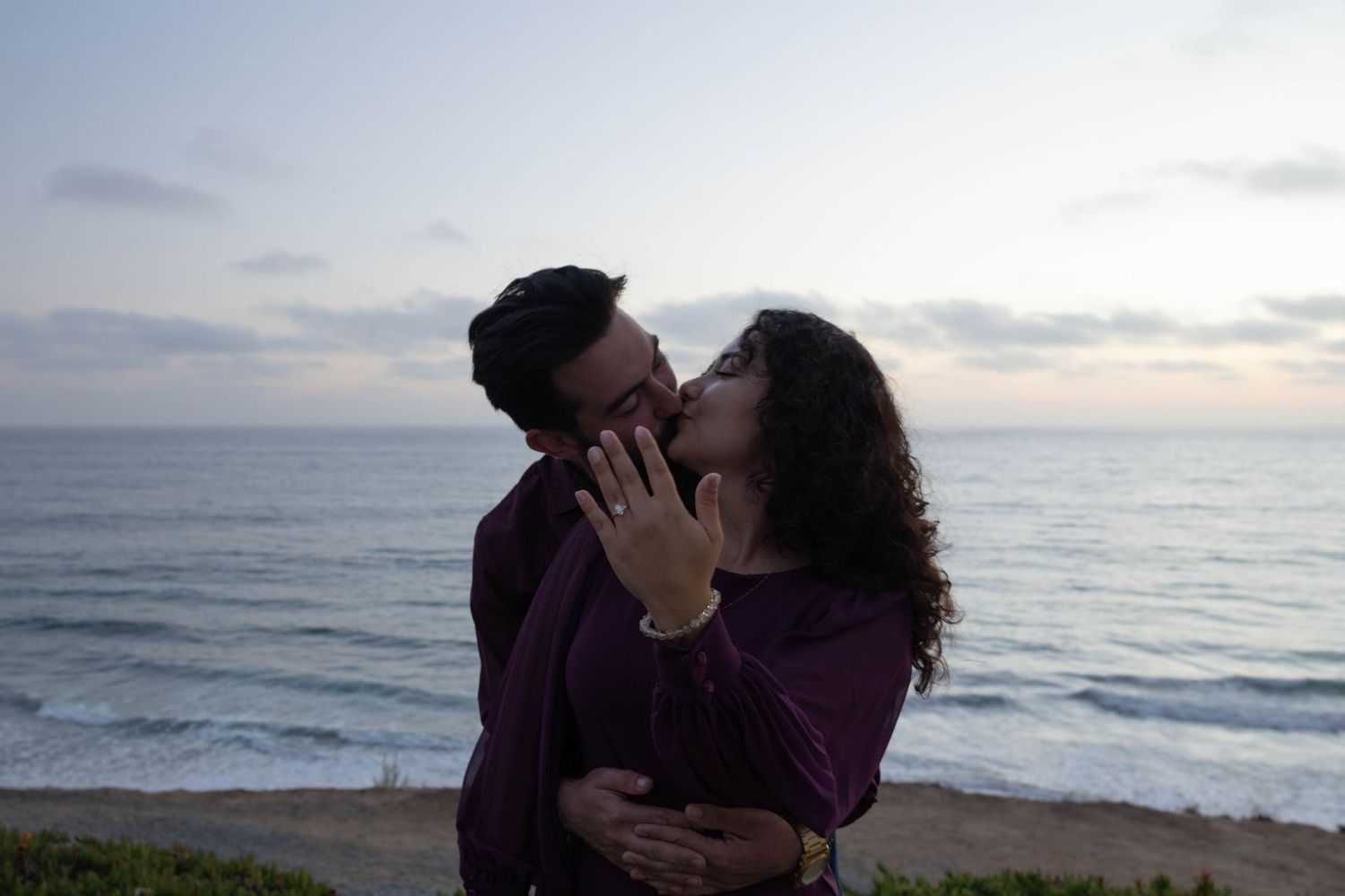 Couple sharing a romantic kiss by the ocean at sunset, both wearing purple, with waves and cloudy sky in background. Beach portraits photos.