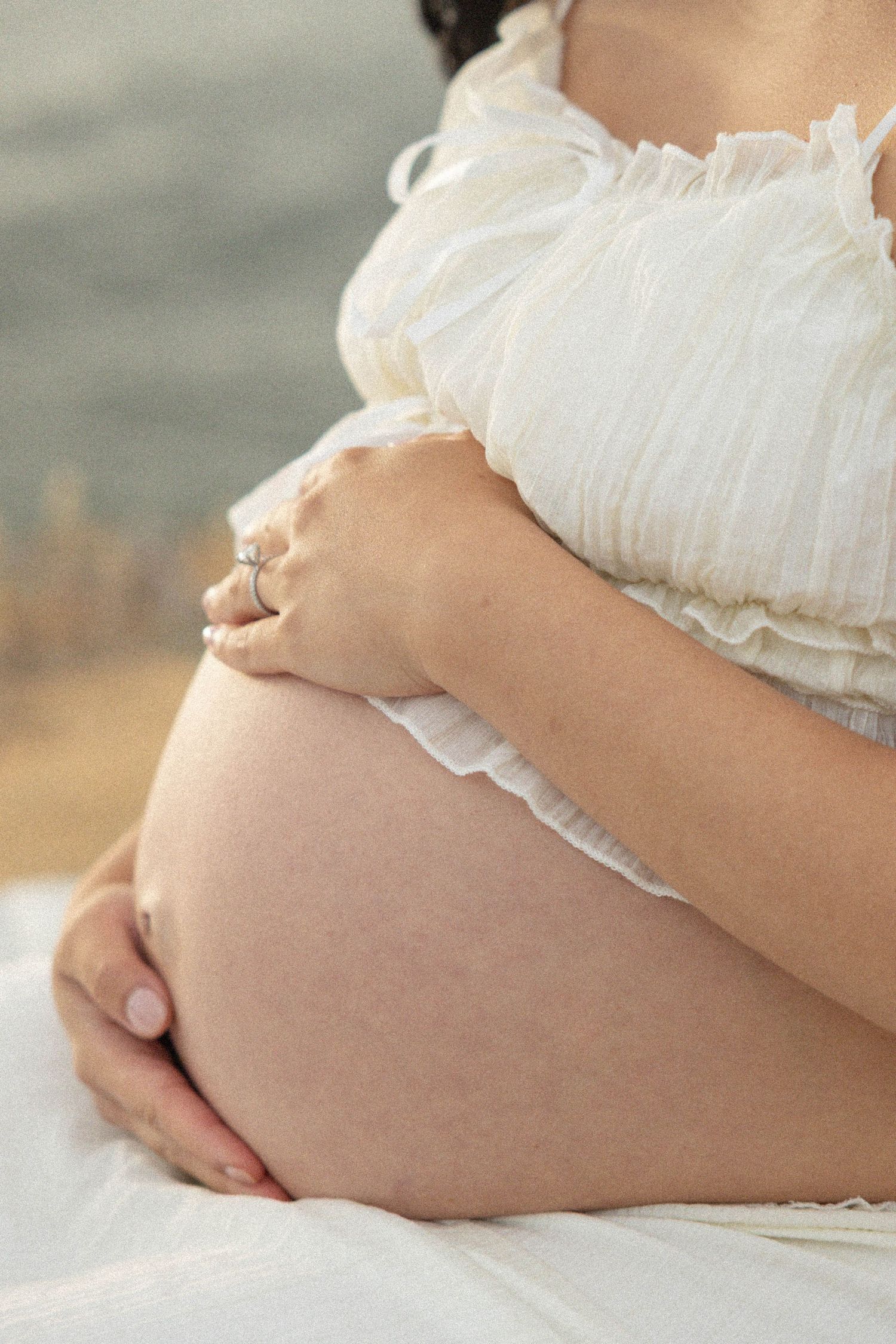 Pregnant woman in white ruffled dress cradling baby bump with ring on finger.