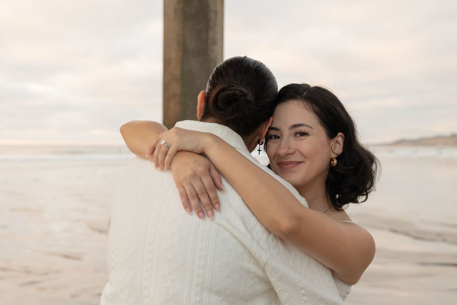 A woman in a white dress smiles while embracing her partner on a beach at sunset. San Diego Engagement Photographer.