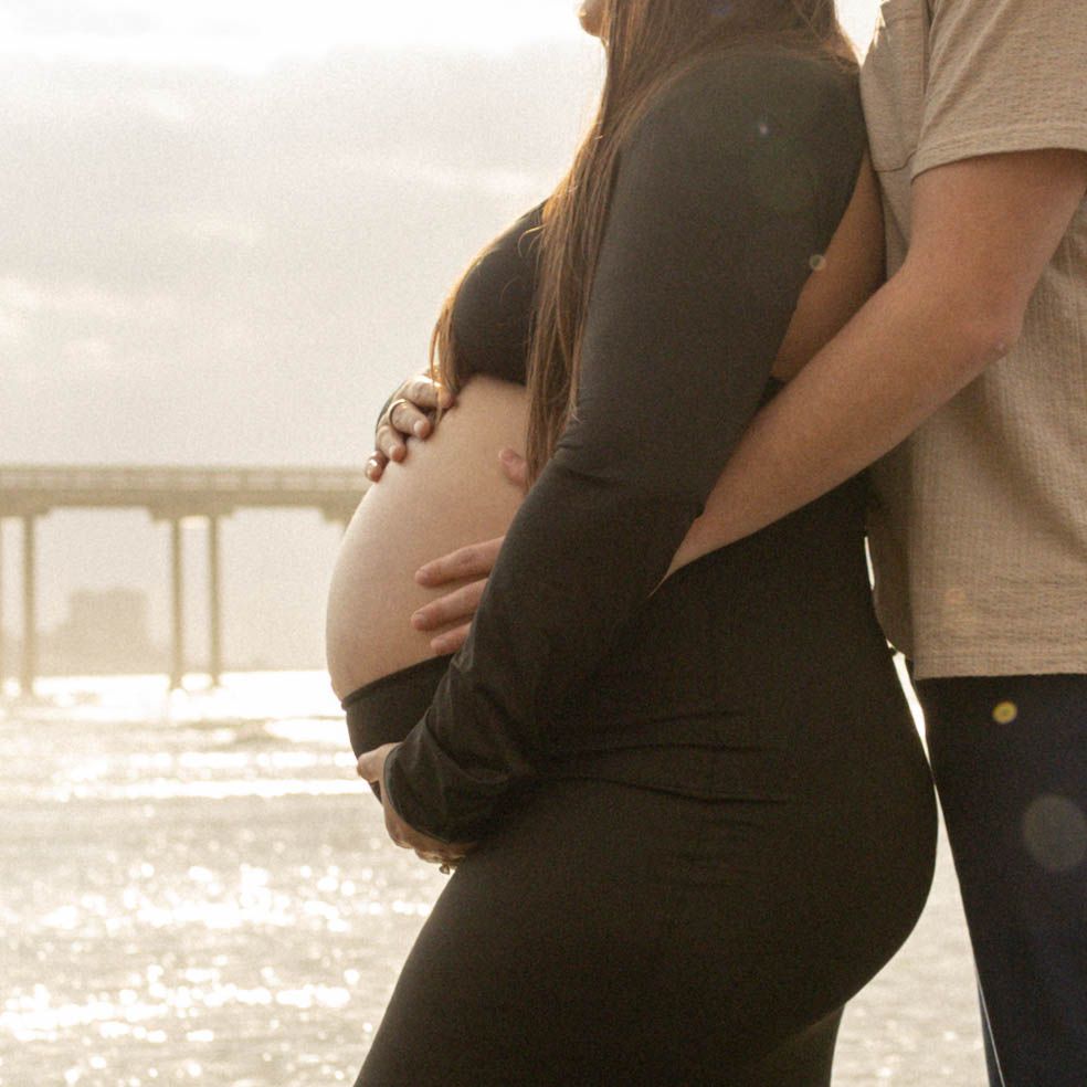 Pregnant couple embracing on a sunlit beach, woman in black dress cradling baby bump near a pier. Maternity Photographer