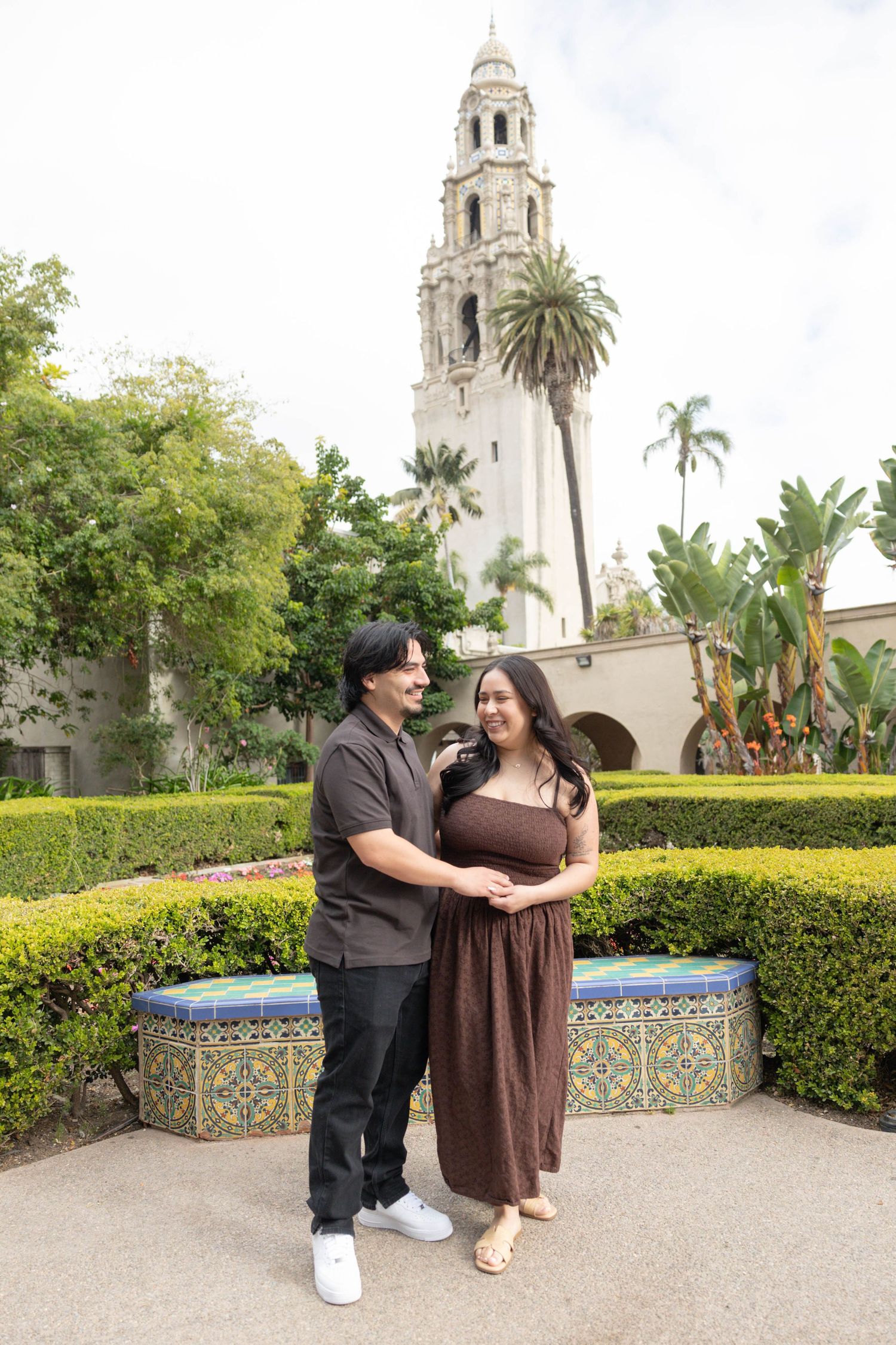 Couple embracing in front of California Tower at Balboa Park, San Diego, surrounded by lush hedges and colorful tile bench. Couples Portrait