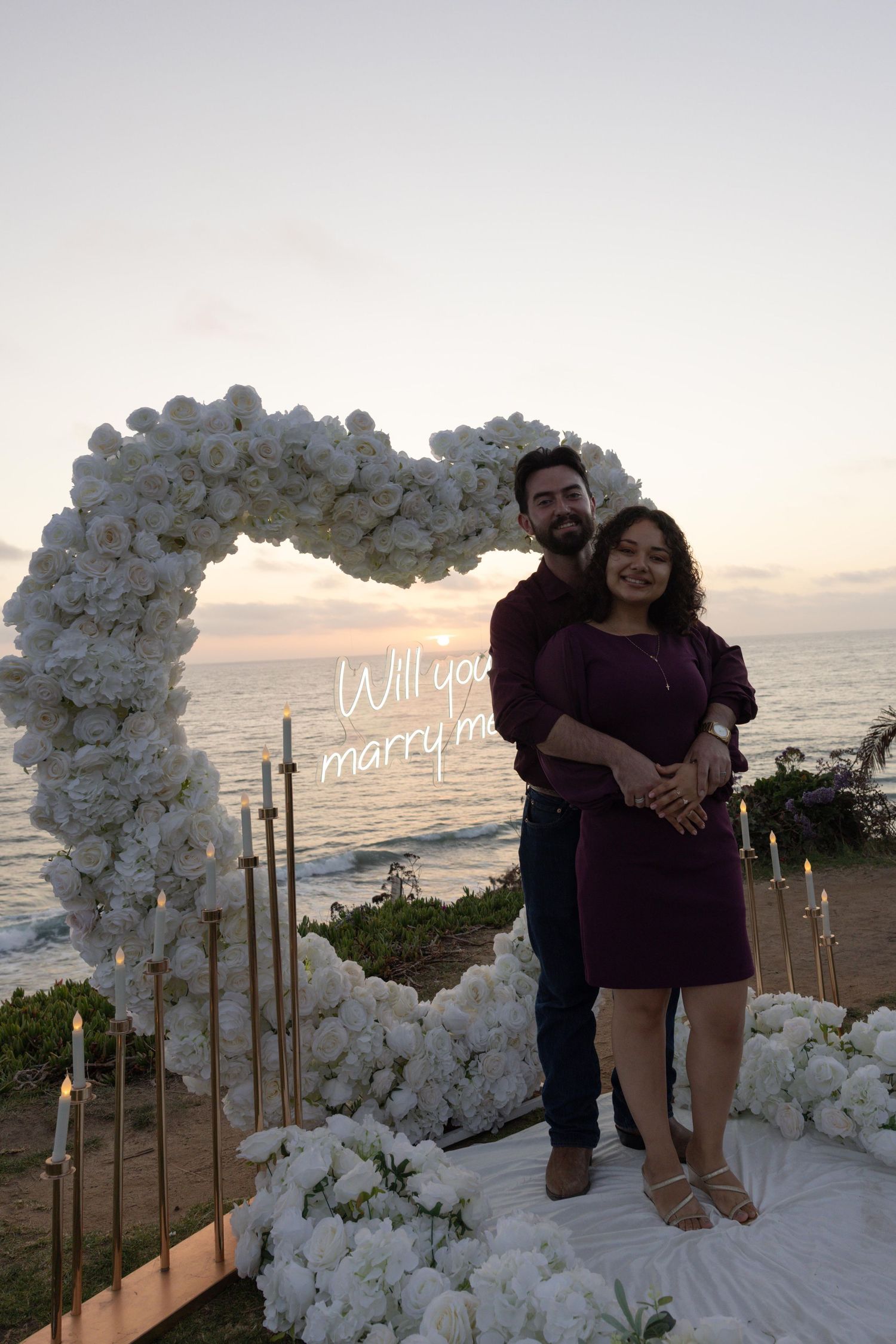 Couple poses under white floral heart arch with Will You Marry Me sign at sunset beach proposal. Del Mar. Southern California Photography.