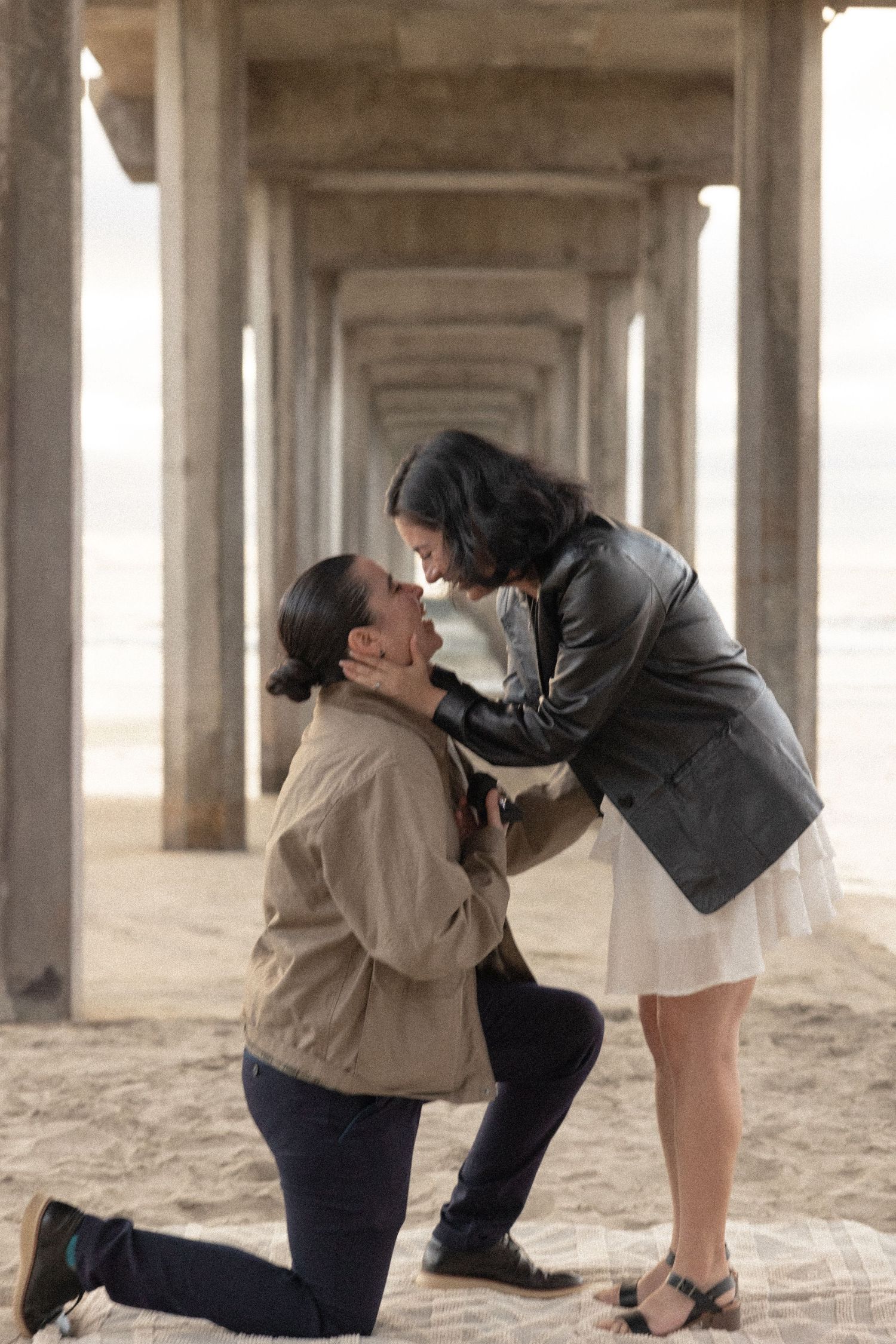 Woman proposing on one knee under a pier on sandy beach as woman leans down to kiss her.