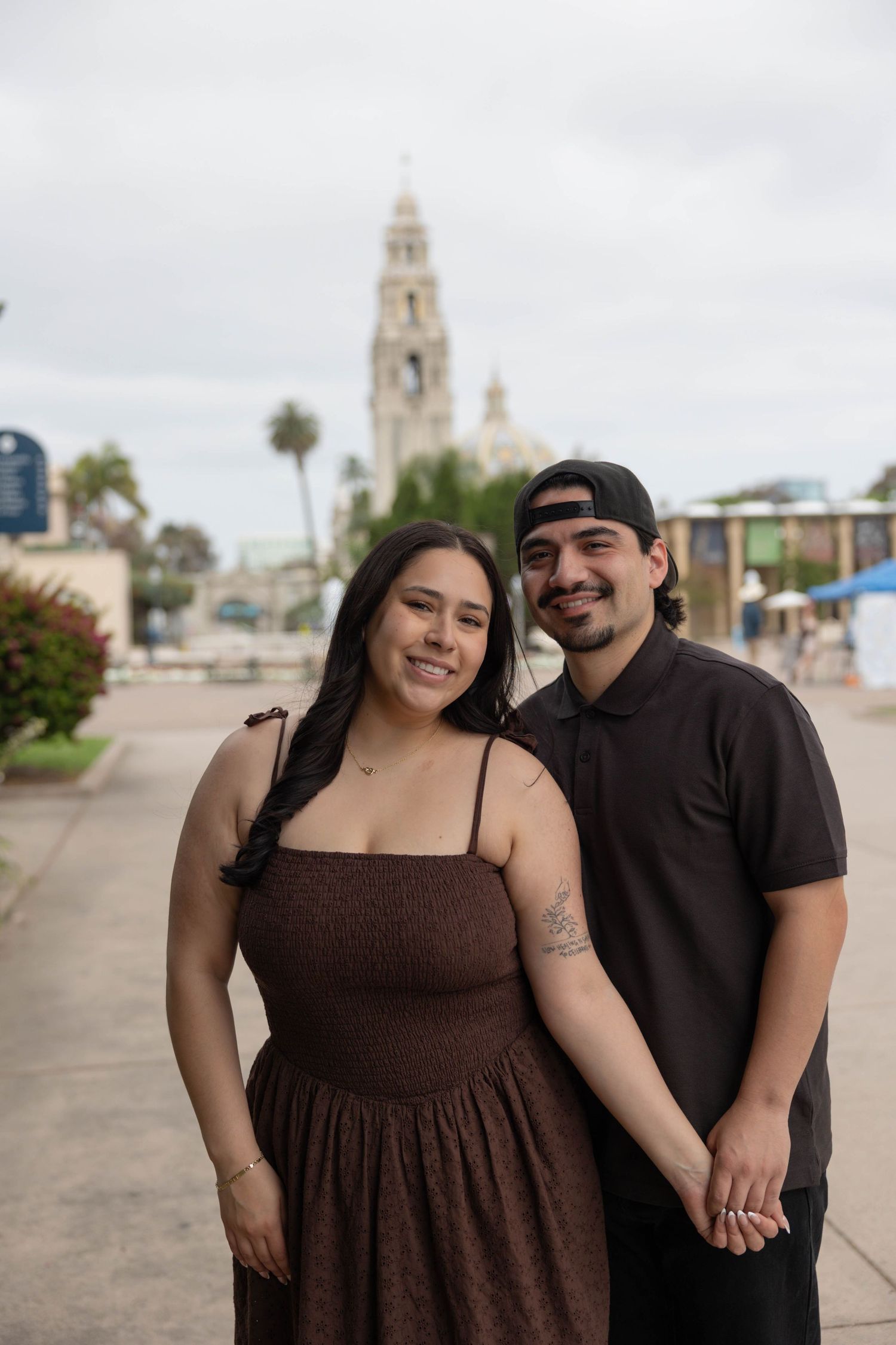 A smiling couple poses together at Balboa Park in San Diego, with the California Tower visible in the background. Couples Portraits.