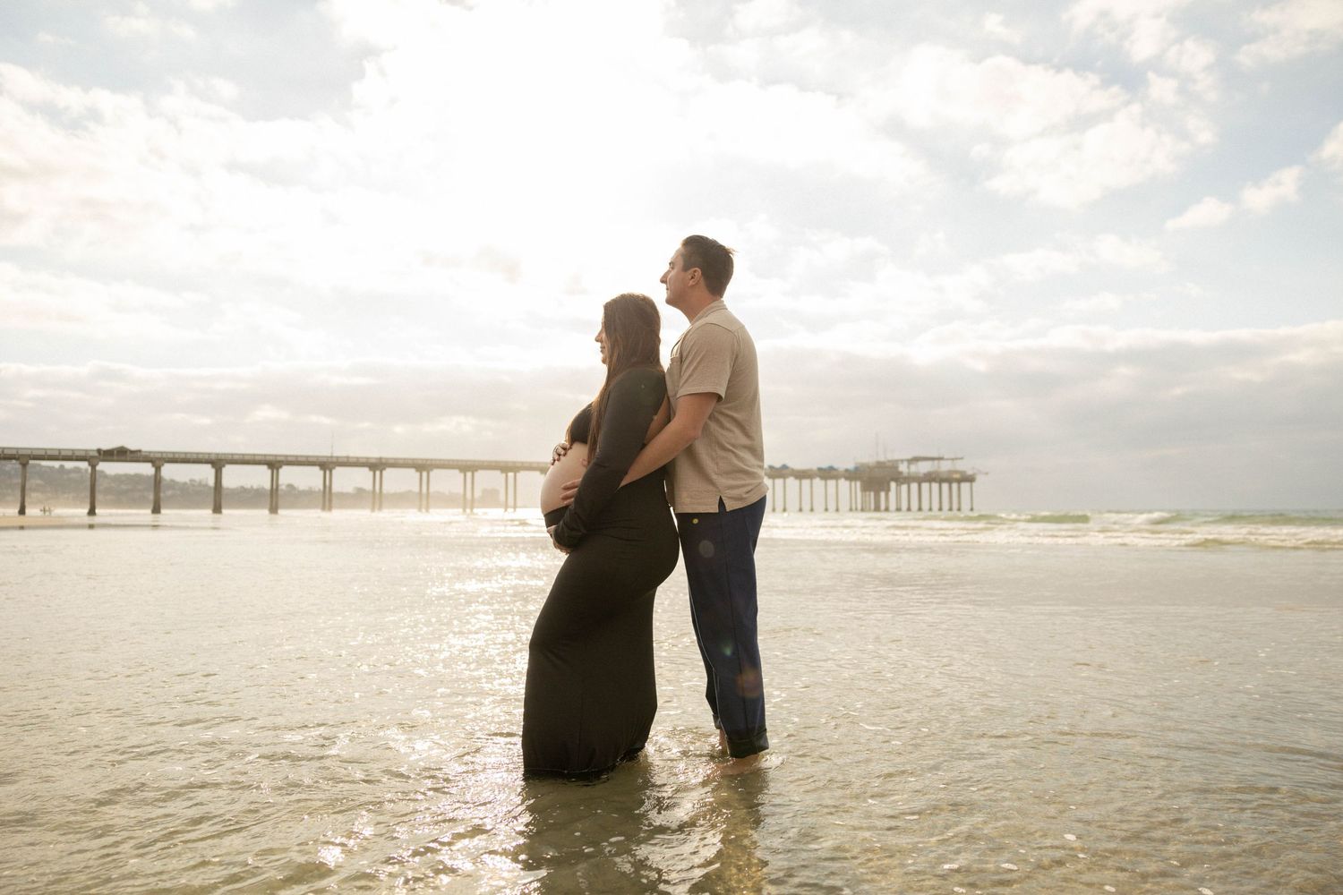 Pregnant couple standing in ocean waves near a pier, sharing a tender moment with soft sunlight glowing behind them. Maternity Photographer.