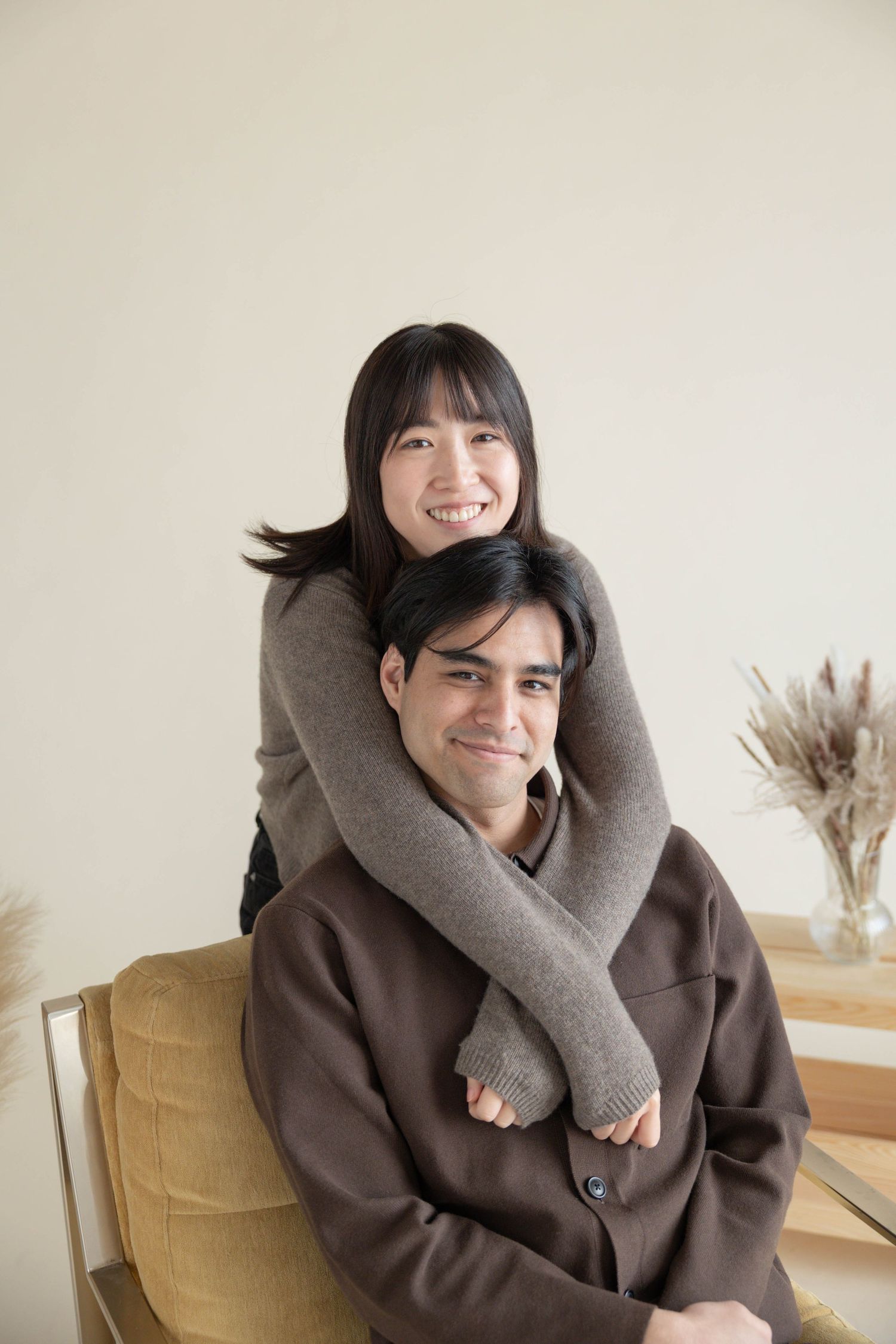 A smiling young Asian couple posing together, woman hugging man from behind on a cozy sofa in a bright room. Couples Portraits San Diego Ca