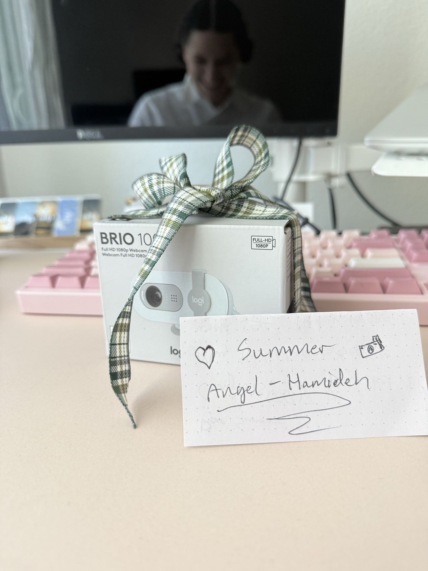 A gift-wrapped Brio 100 webcam with plaid ribbon bow and a handwritten note reading Summer Angel-Hamideh on a desk.