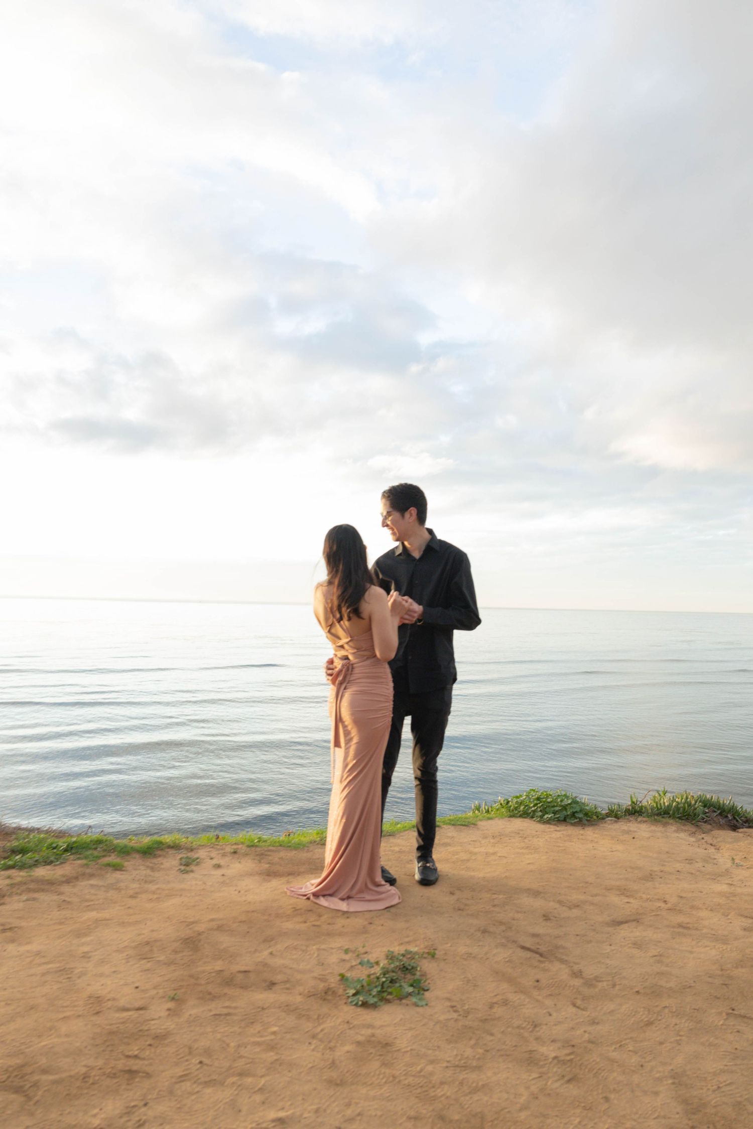 Couple in formal attire embracing on a coastal cliff overlooking a calm ocean at sunset. San Diego Engagement Photographer. Sunset Cliffs.