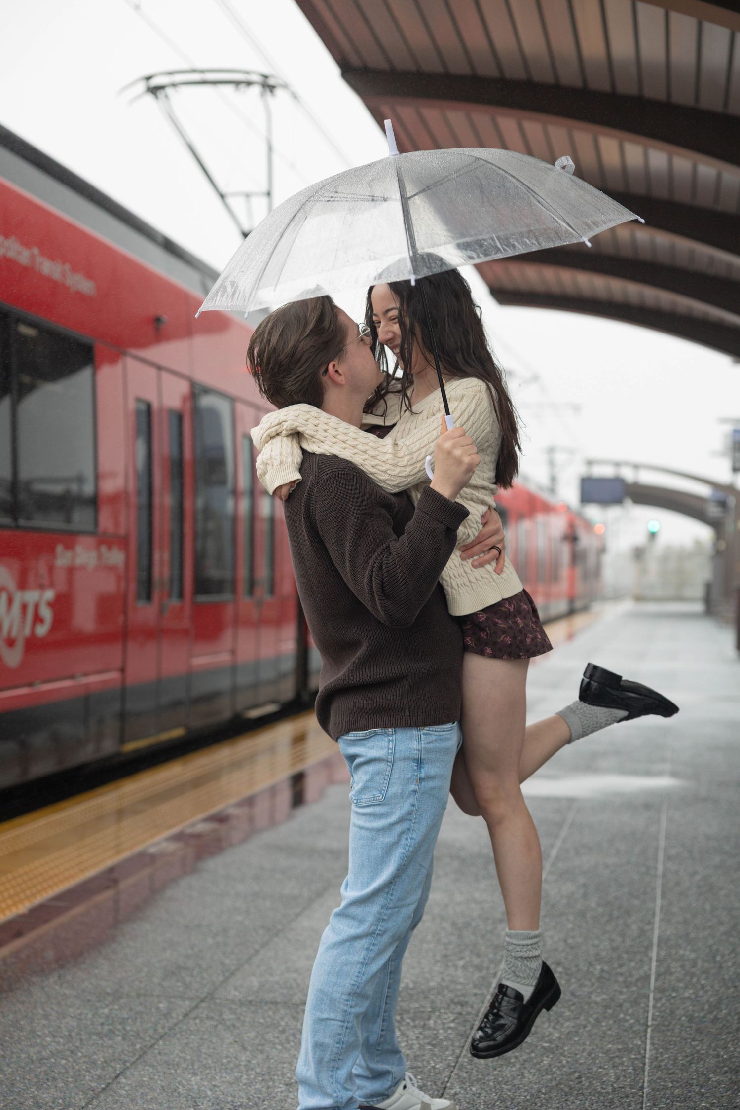 Male holding Female up in the air as they kiss under a transparent umbrella at the MTS trolley stop in San Diego.