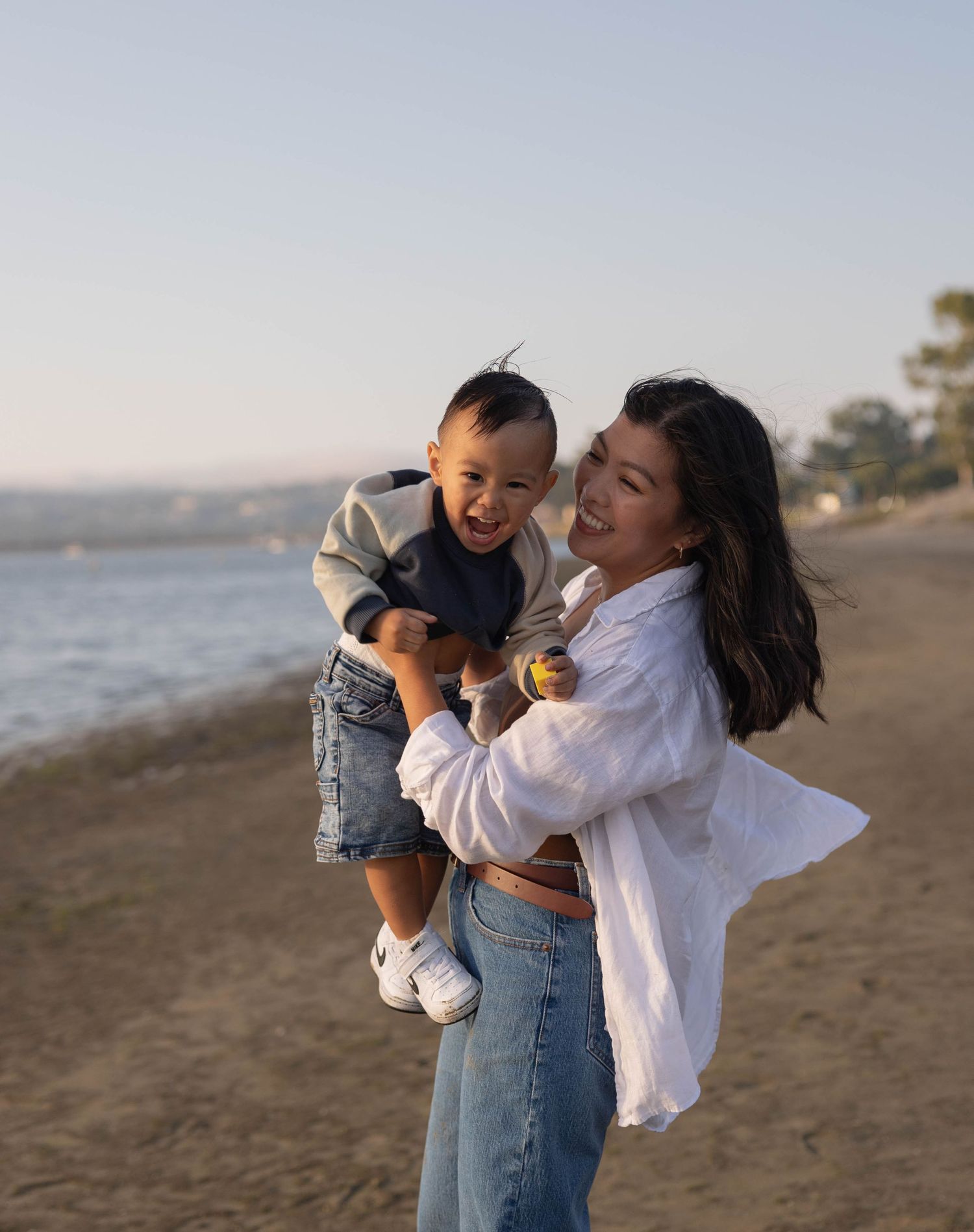 A woman in a white shirt and jeans laughs while holding a happy toddler on a sunny beach. Family Photographer.