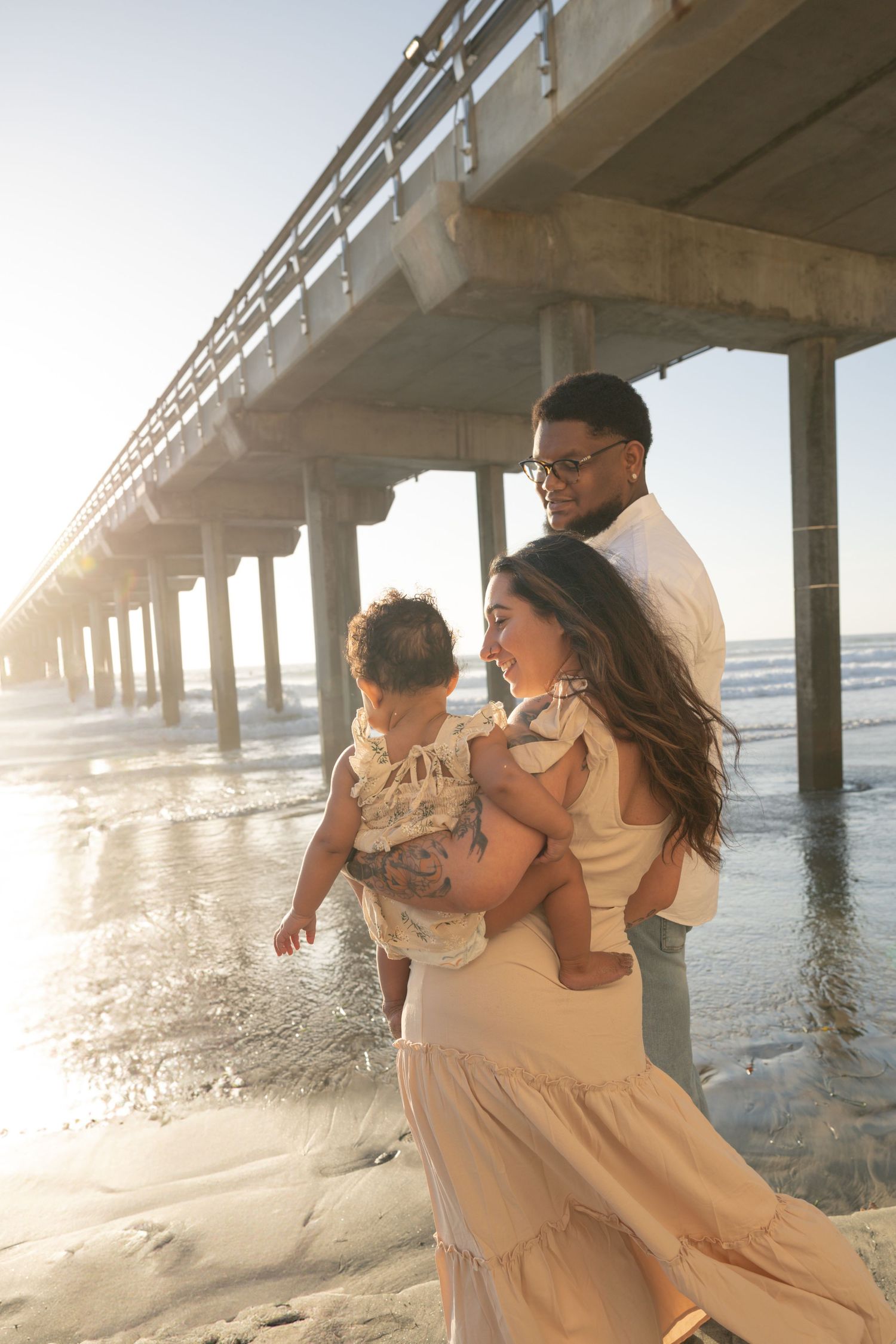 Family of three poses near a sunlit beach pier, mother holding baby in cream outfits while father stands behind them. Maternity Photographer