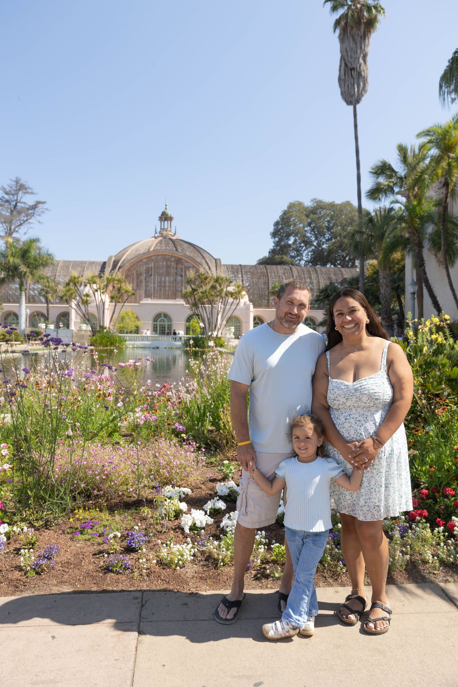 Family of three posing in front of a botanical garden with colorful flowers and a domed greenhouse on a sunny day.