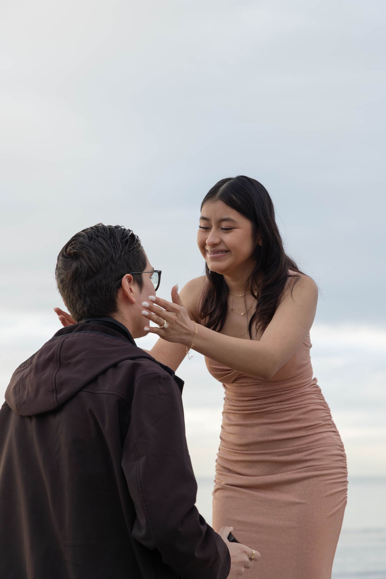 A woman in a pink dress smiles while touching a man's face near the ocean during a romantic proposal moment. San Diego Engagement Photograph
