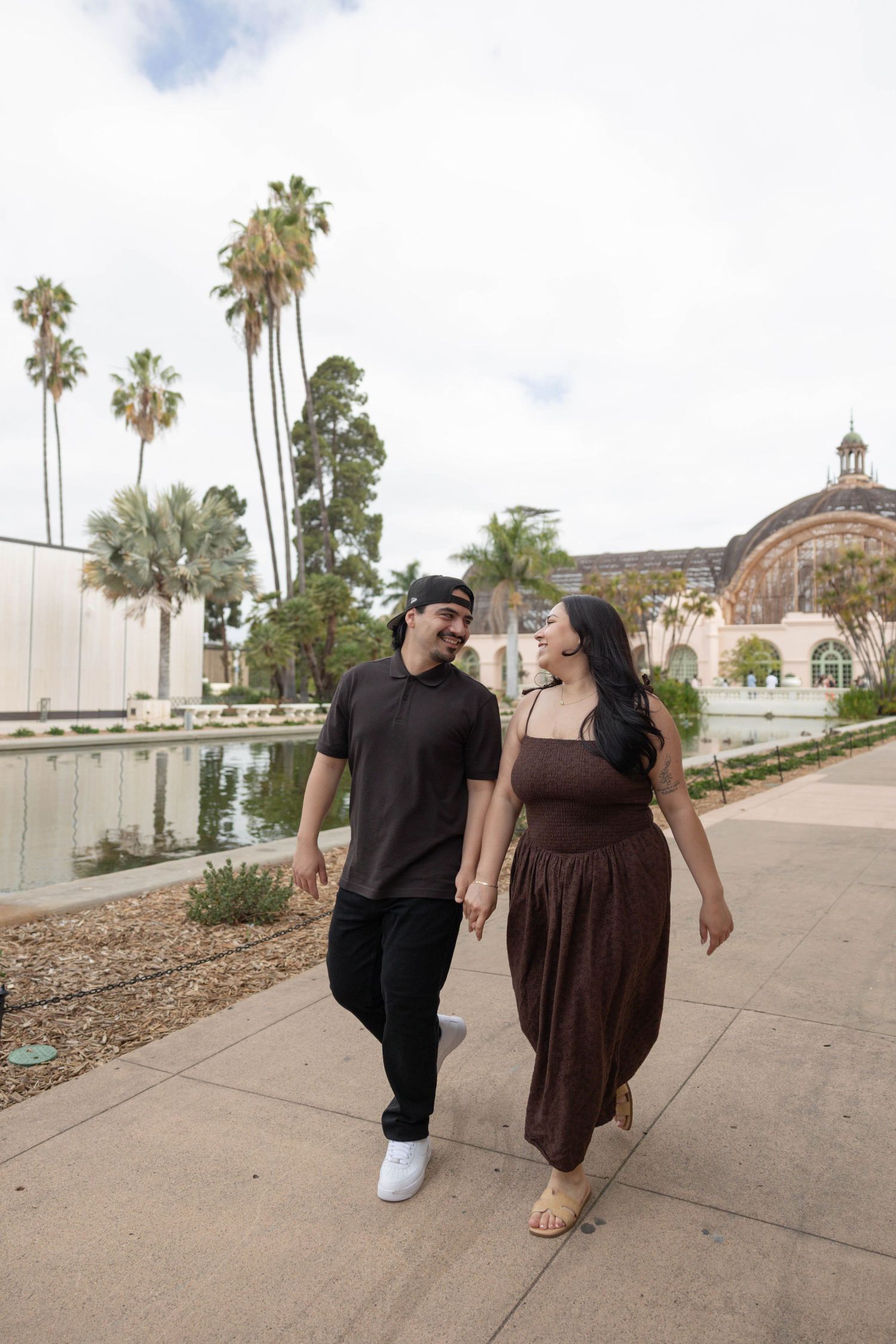 Couple holding hands and walking along a sunny path at Balboa Park, San Diego, with palm trees and historic building. Couples Portraits SD