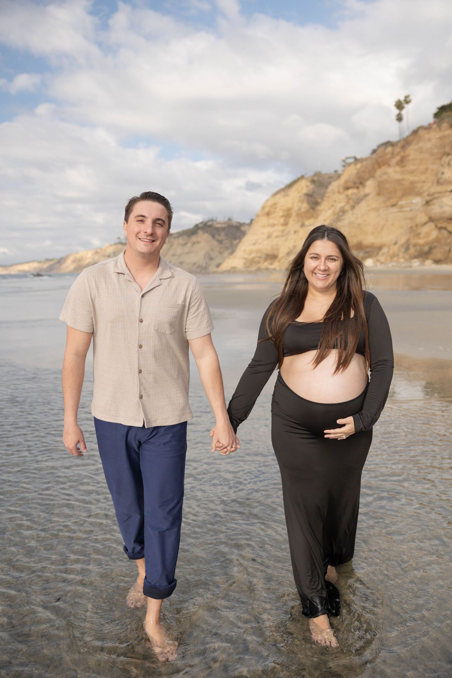 Pregnant couple holding hands walking barefoot on a beach with rocky cliffs, woman in black dress cradling baby bump. La Jolla Shores Photos