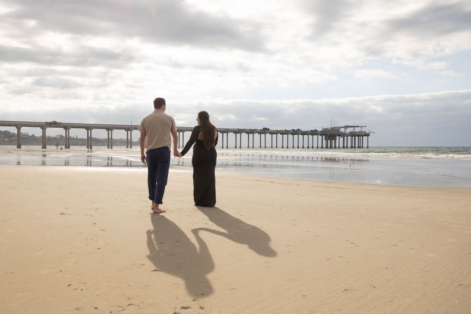 Couple walking hand in hand on a sandy beach toward an ocean pier under overcast skies.