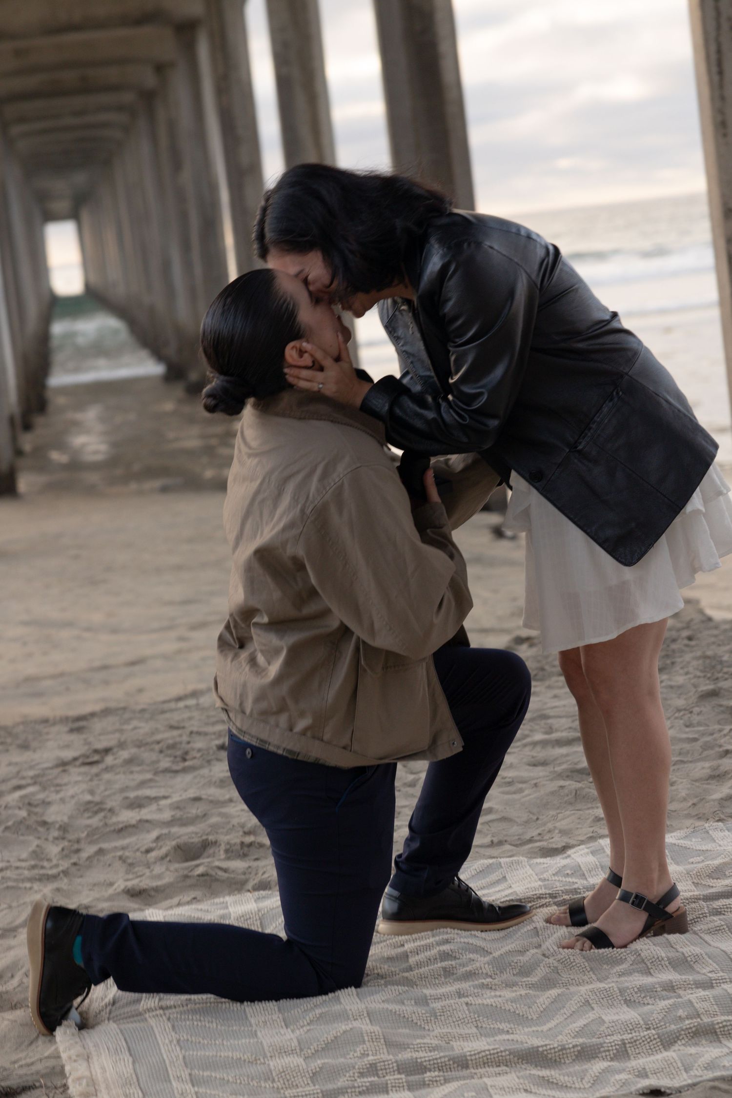 Couple sharing a romantic kiss during a beach proposal, man on one knee on sandy shore near pier. San Diego Engagement Photographer.