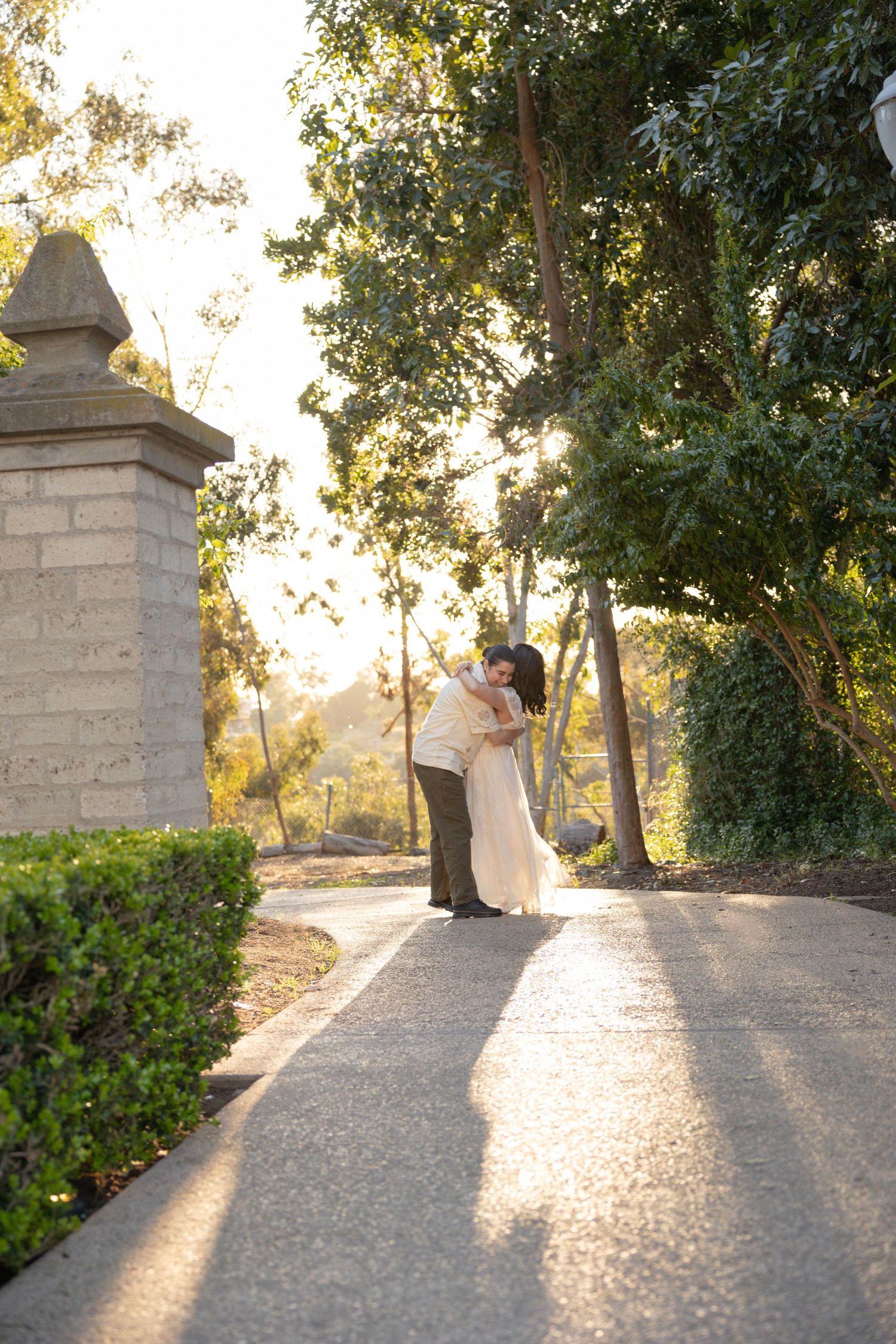 Couple embracing on a sunlit garden pathway surrounded by lush trees and hedges during golden hour. Couples Portraits San Diego California