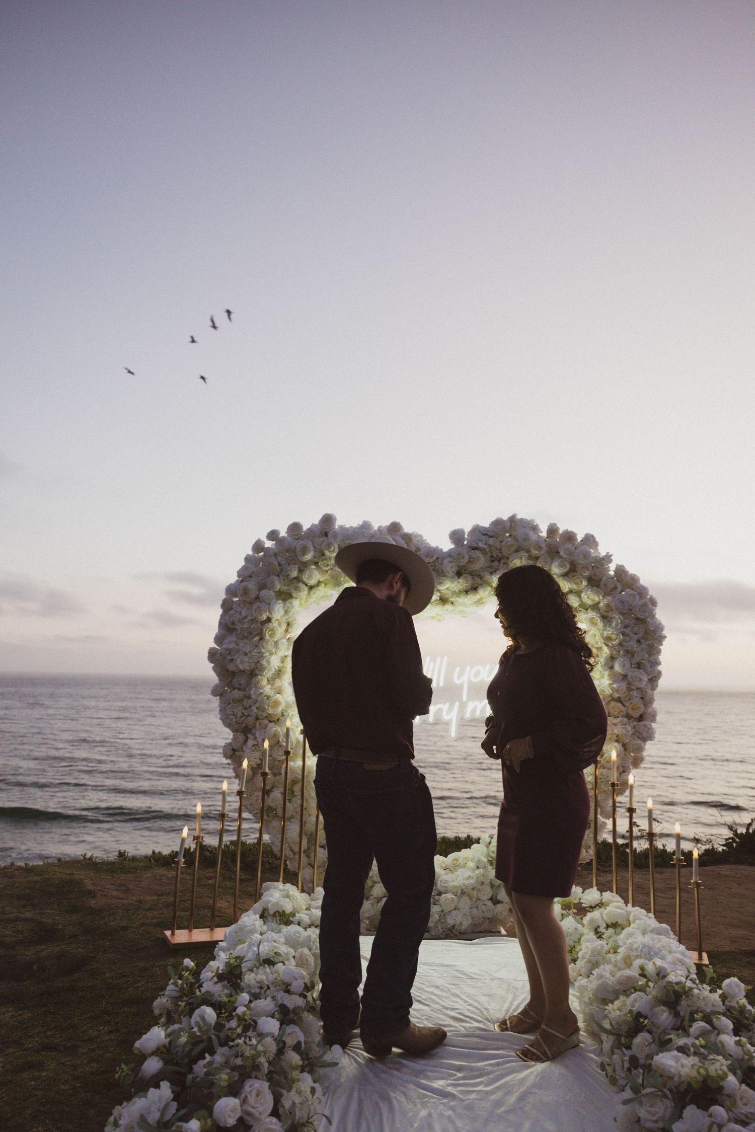 Couple silhouetted before heart-shaped floral arch with candles by the ocean at dusk. Engagement Photos.