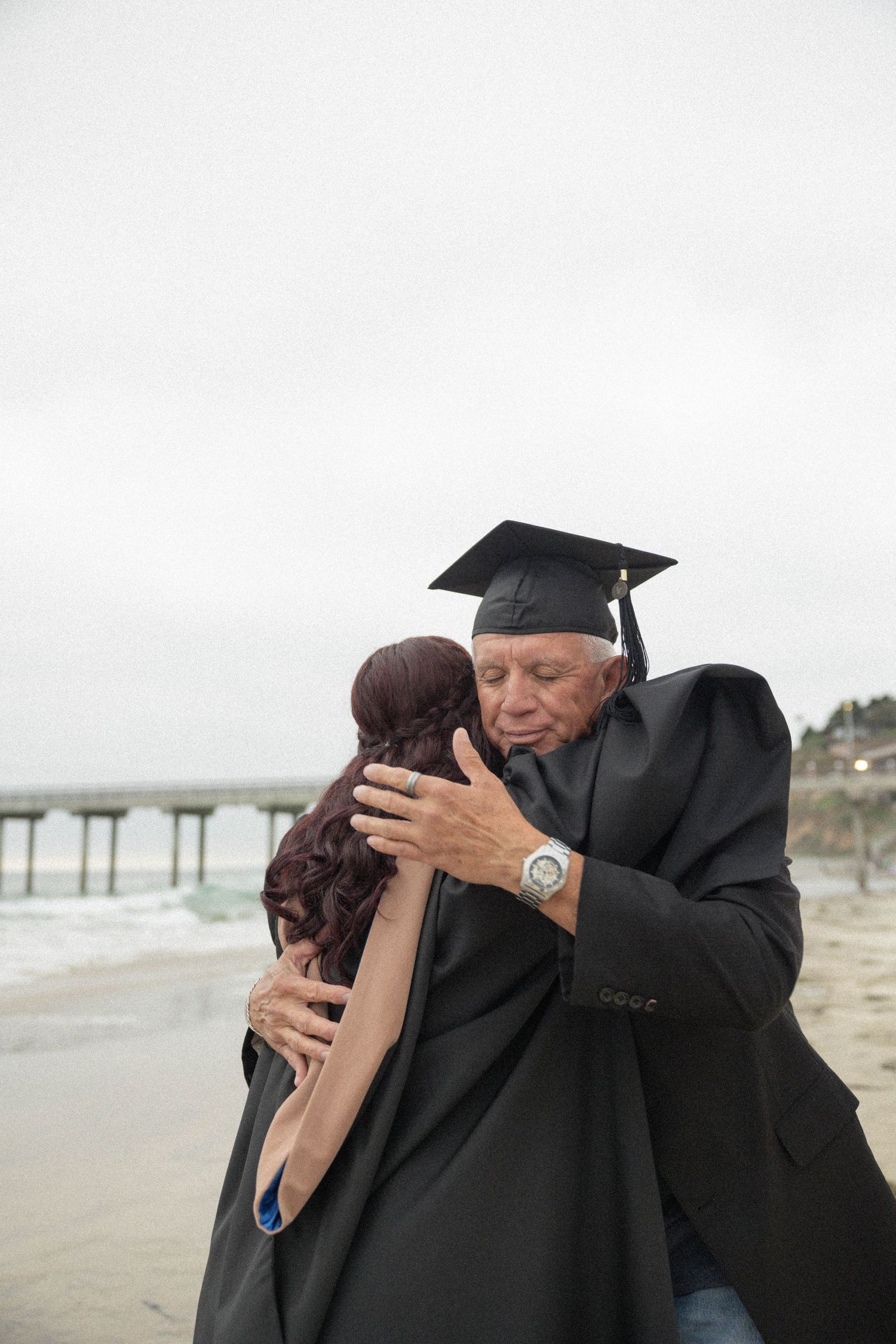 Graduate in cap and gown sharing emotional hug on beach near pier.