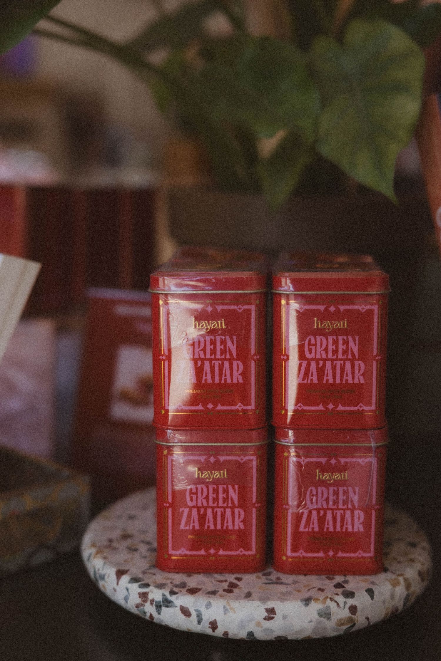 Four red tins of Green Zaatar spice stacked on marble surface with plant in background.