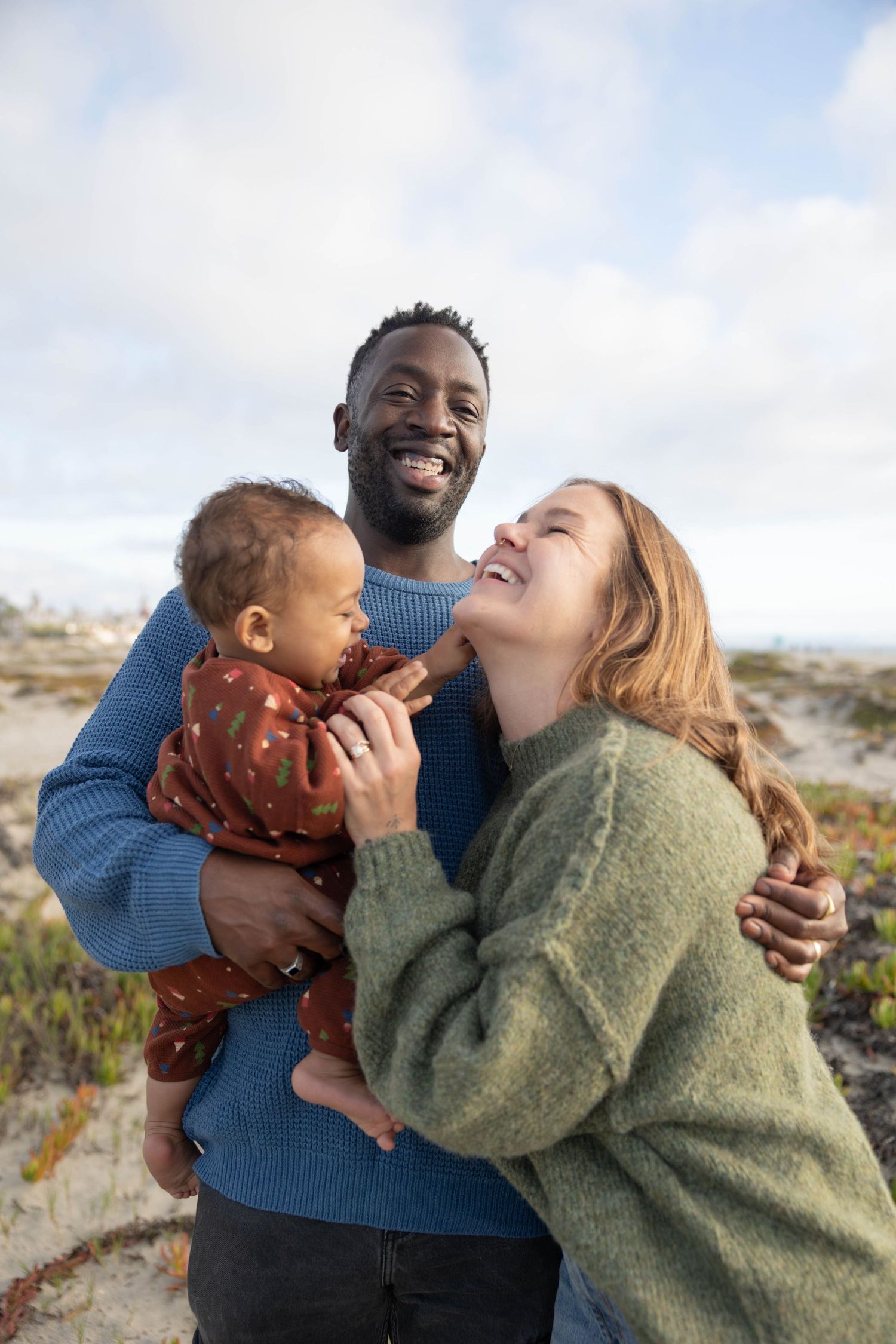 Happy interracial family with mother, father, and baby smiling outdoors at the beach on a cloudy day.