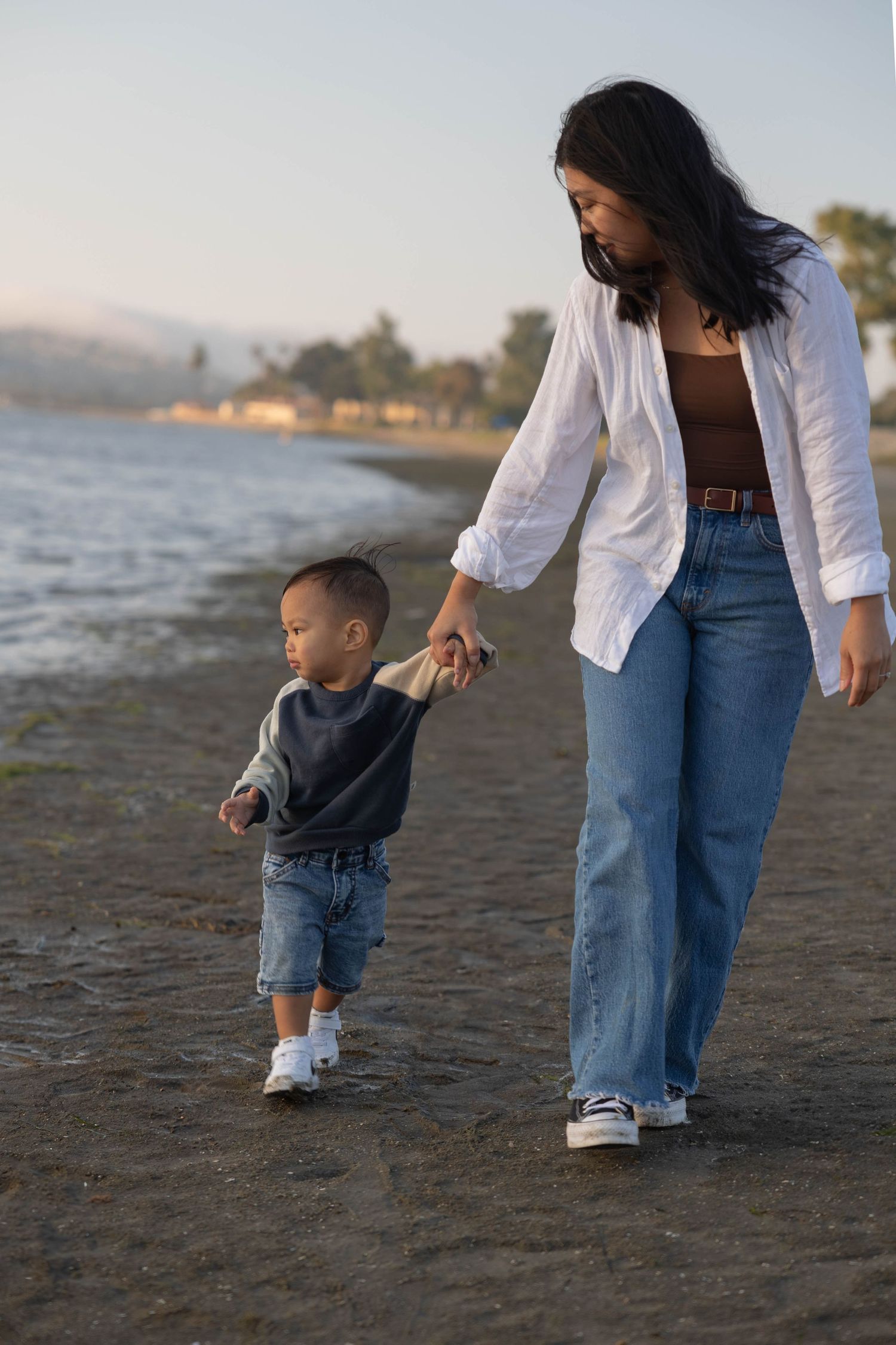 A mother and young son walk hand-in-hand along a sandy beach at sunset, both wearing casual clothing. Family Photographer.