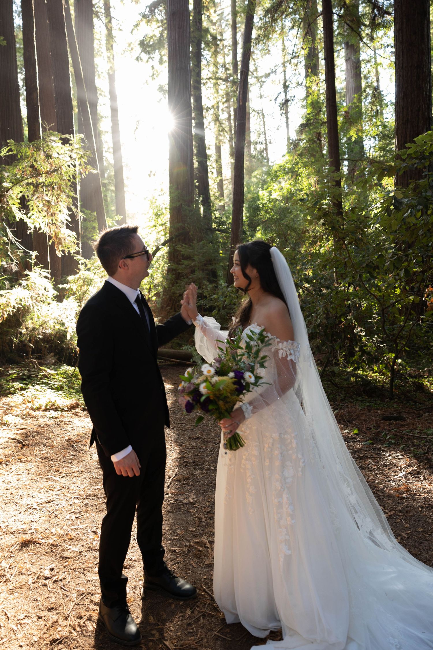 Bride and groom share a joyful first look moment in a sunlit redwood forest, bride holding a lush floral bouquet. Wedding photography.