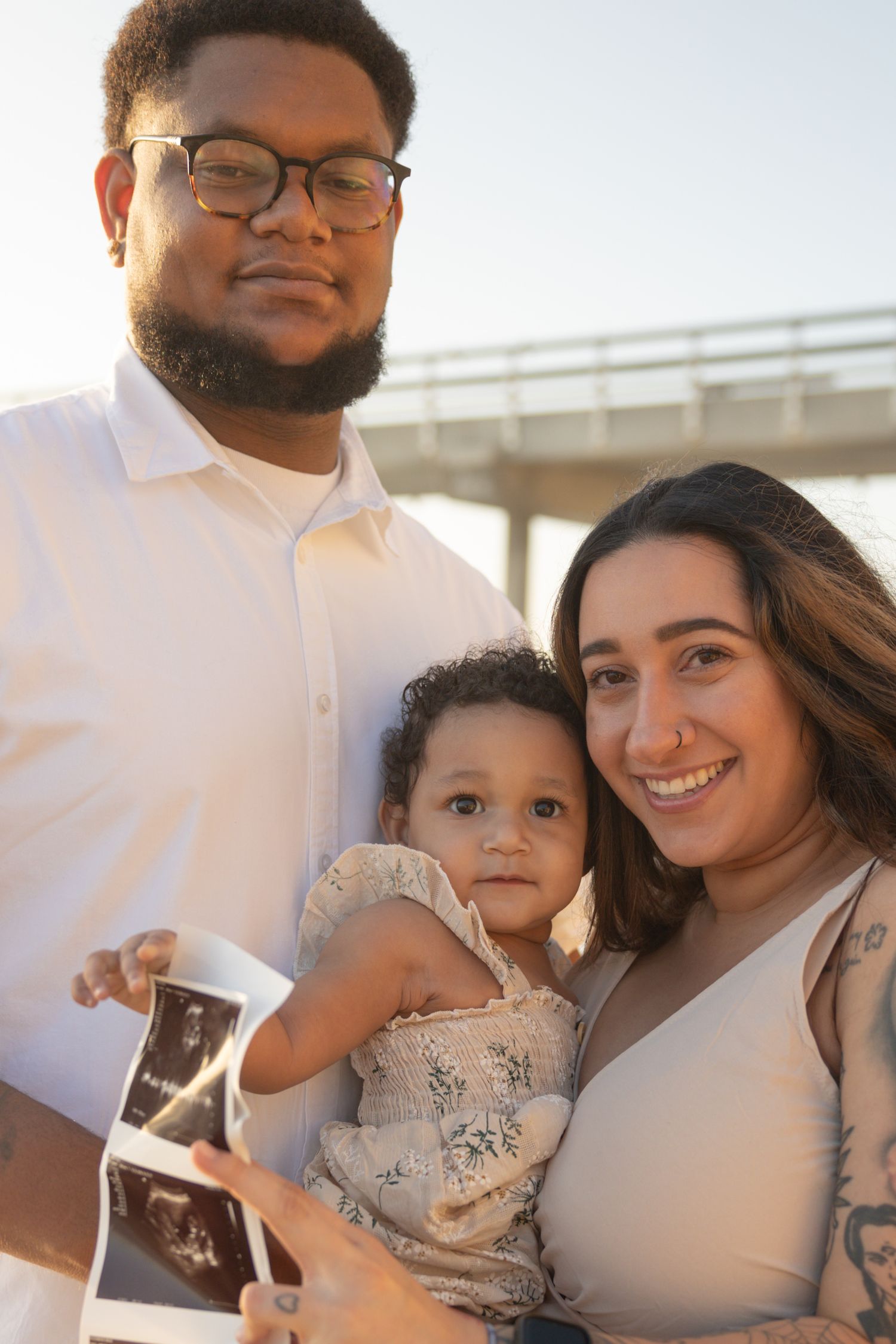Family holding ultrasound photos and toddler at beach pier during golden hour sunset announcement. Maternity Photographer San Diego Ca.