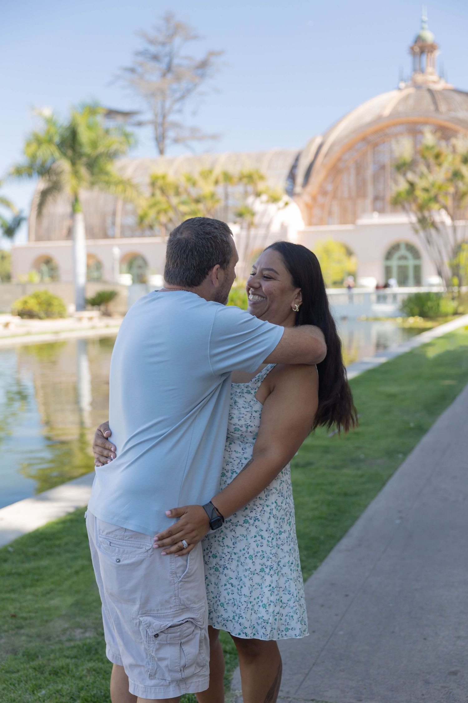 Couple embracing and laughing by a pond at Balboa Park, San Diego, with a domed building and palm trees.