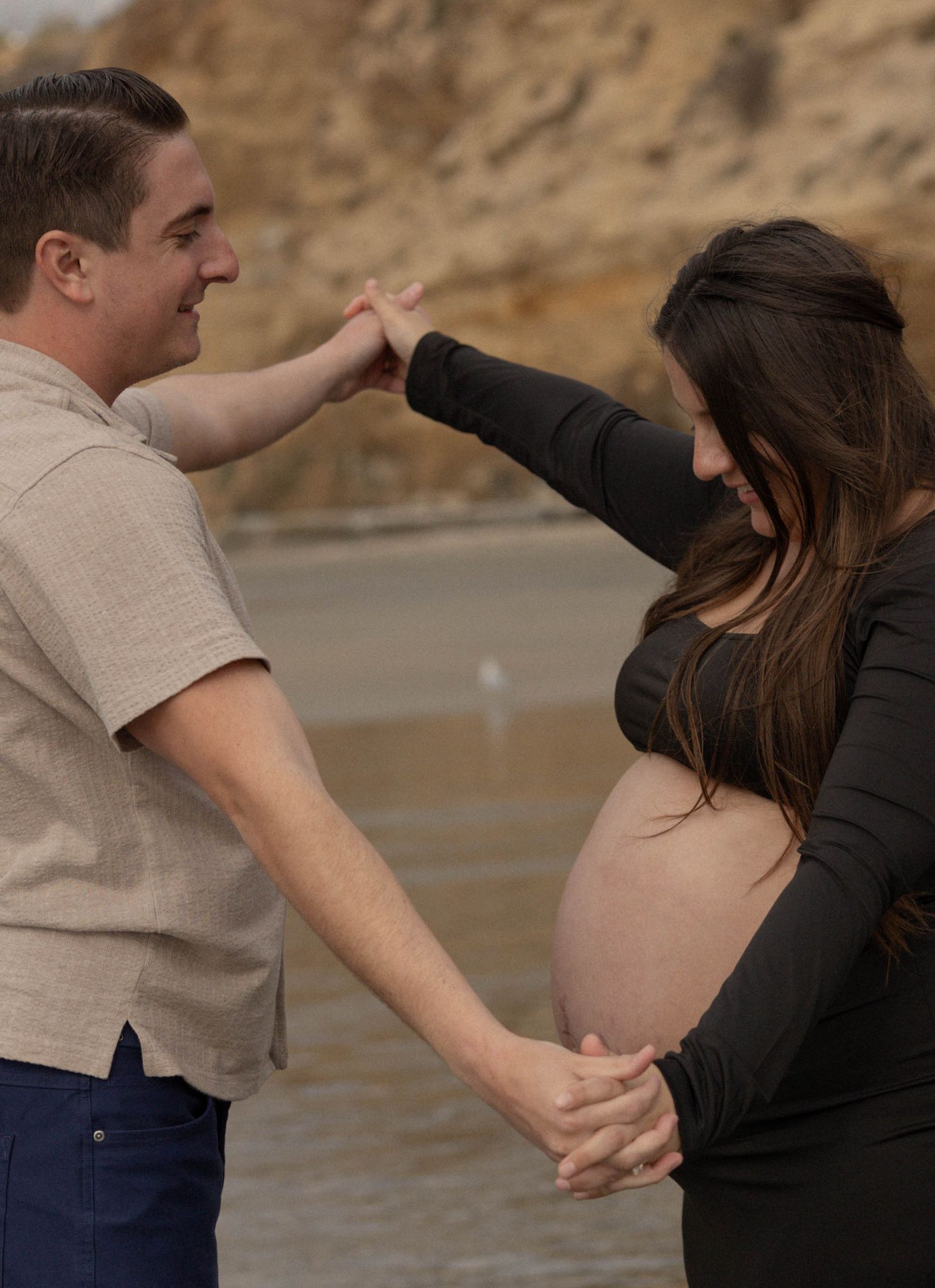 Couple holding hands on a beach, with the pregnant woman cradling her baby bump in a tender maternity photo session. Maternity Photographer.