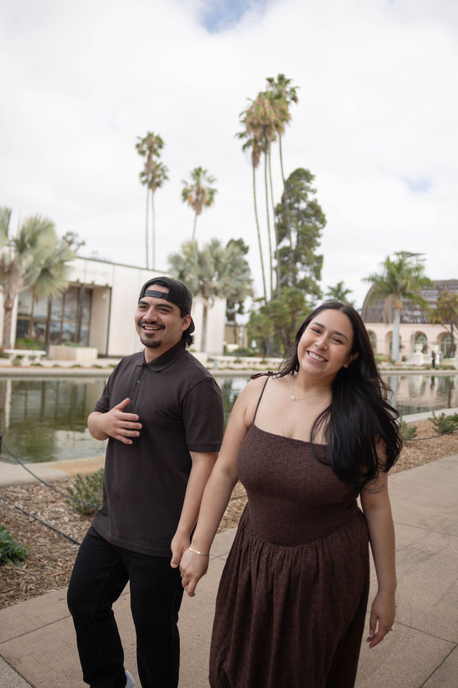 A smiling couple walks hand-in-hand near a pond with tall palm trees in the background on a cloudy day. Couples Portraits San Diego Ca