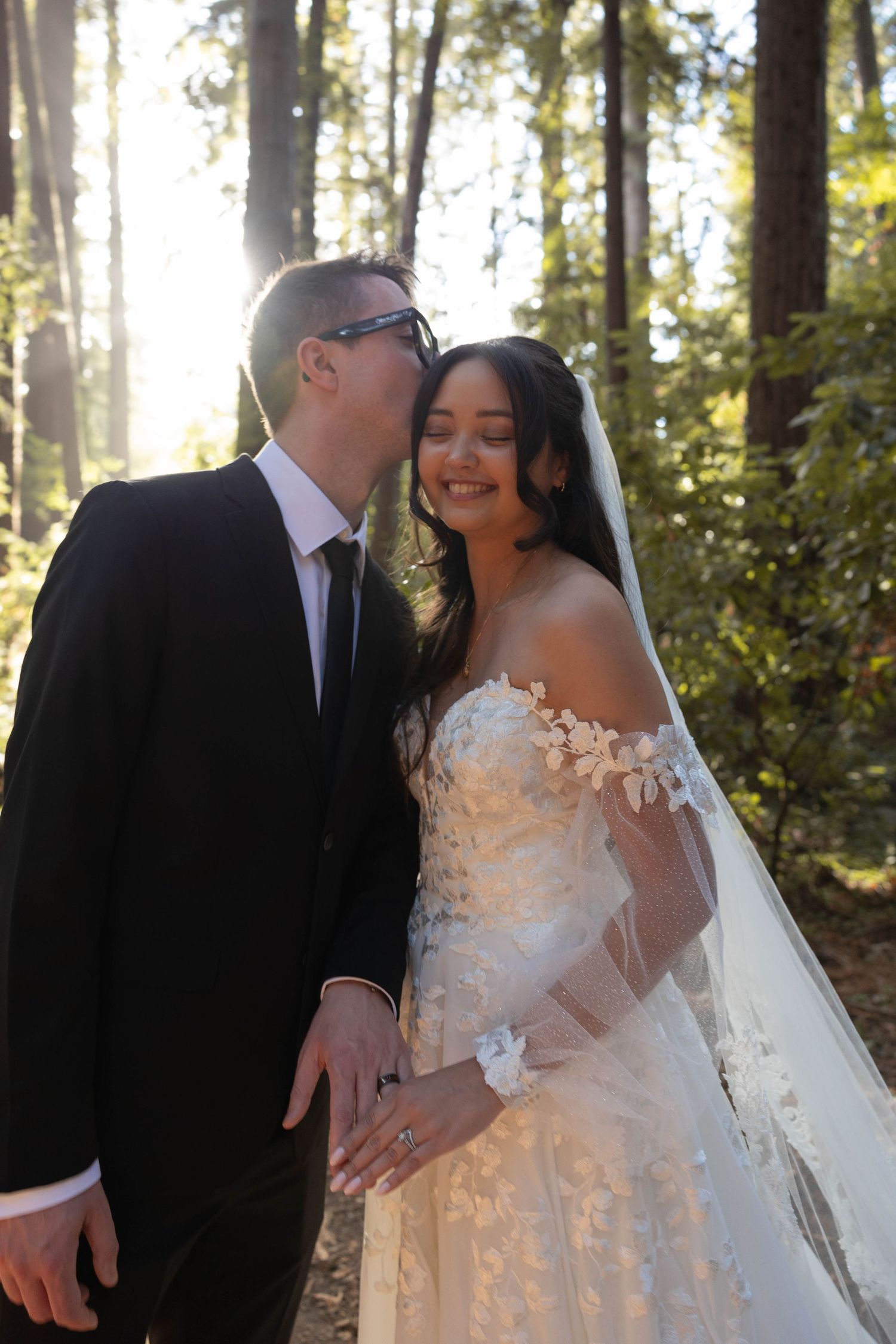 Groom in black suit kissing bride in floral lace off-shoulder gown and veil in sunlit forest wedding portrait. Elopement Photographer.