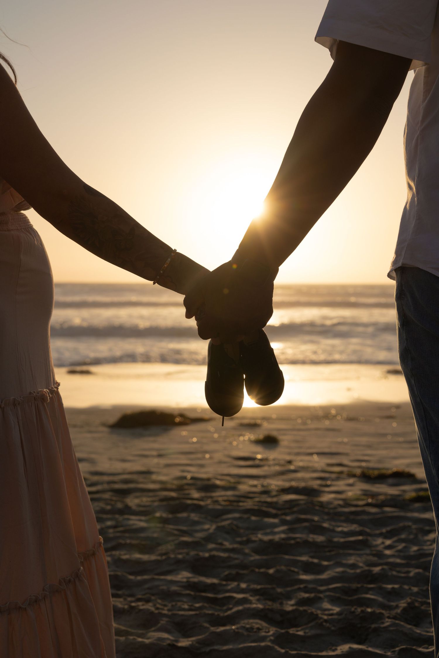 Couple holding hands on a sandy beach at sunset with ocean waves in the background. Sunset. Maternity Photographer San Diego California.