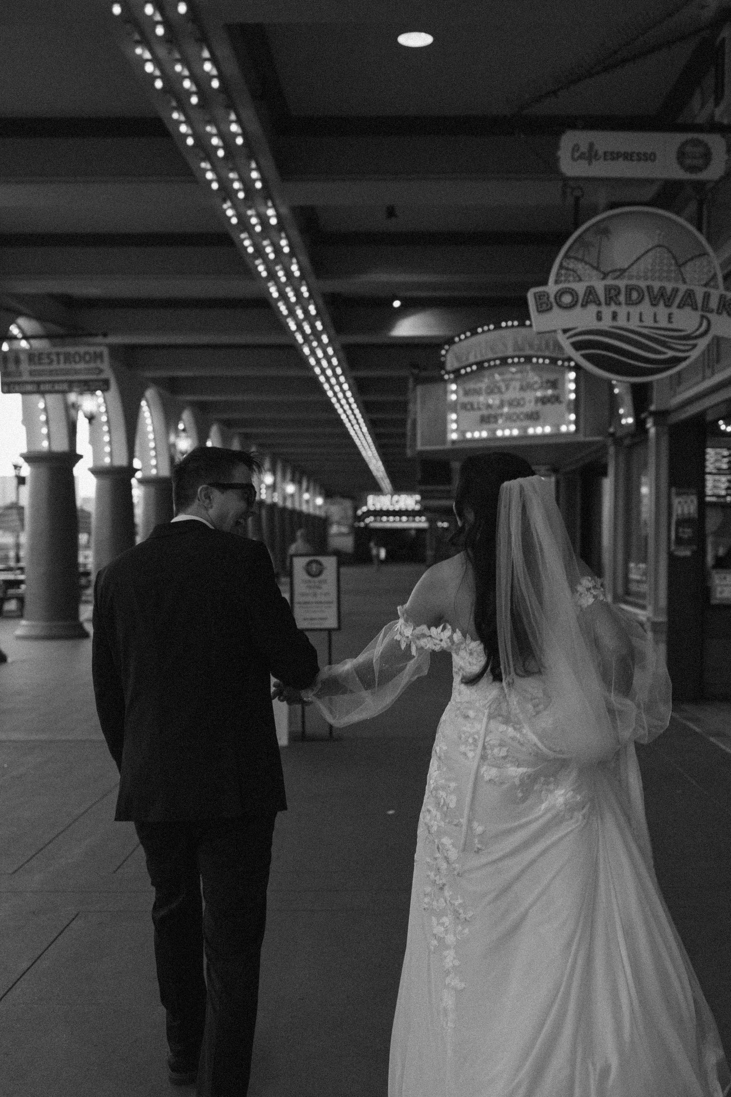 Black and white photo of a bride and groom walking hand in hand past Boardwalk Grille restaurant at night.