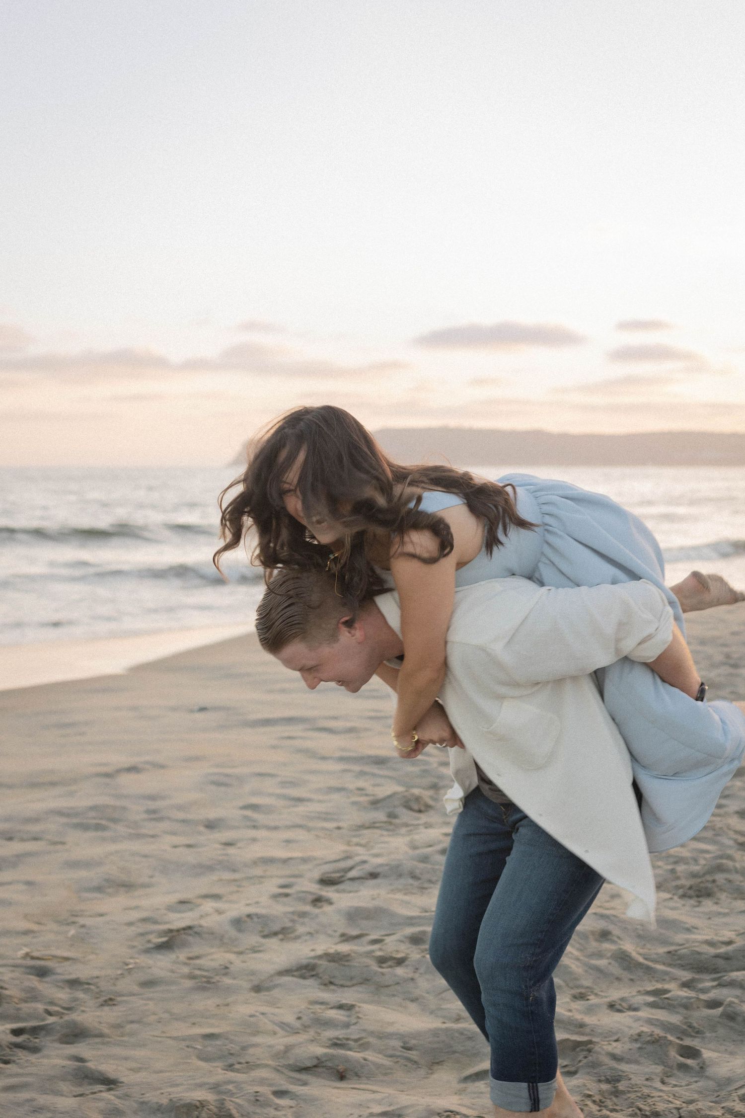 A couple playfully enjoys a piggyback ride on a sandy beach at sunset, with ocean waves in the background.