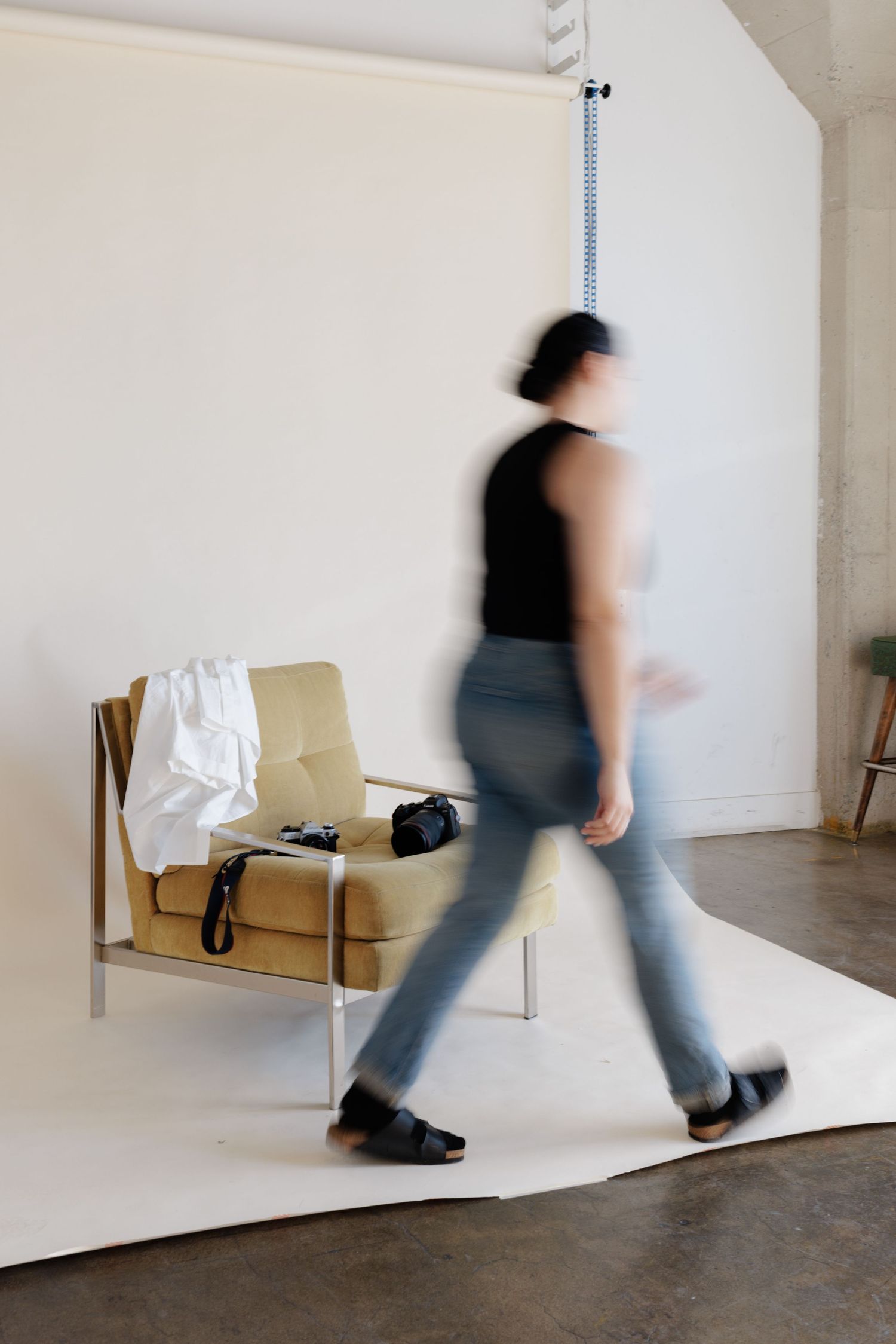 Blurred motion of a person walking past a yellow chair with bags in a minimalist white studio space.